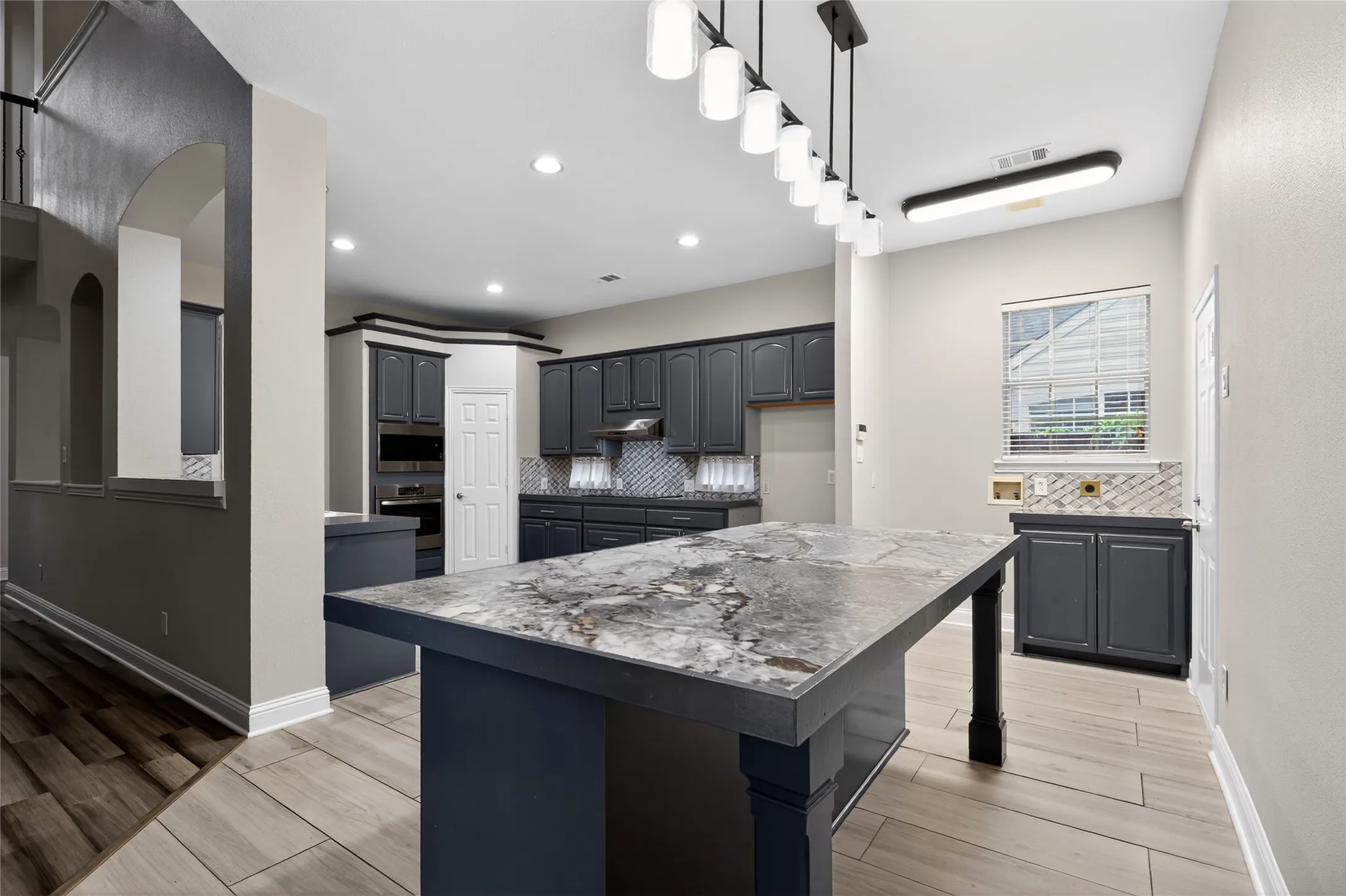 Spacious kitchen featuring an expansive island with a stone-finish countertop, dark cabinetry, herringbone-patterned backsplash, and recessed lighting