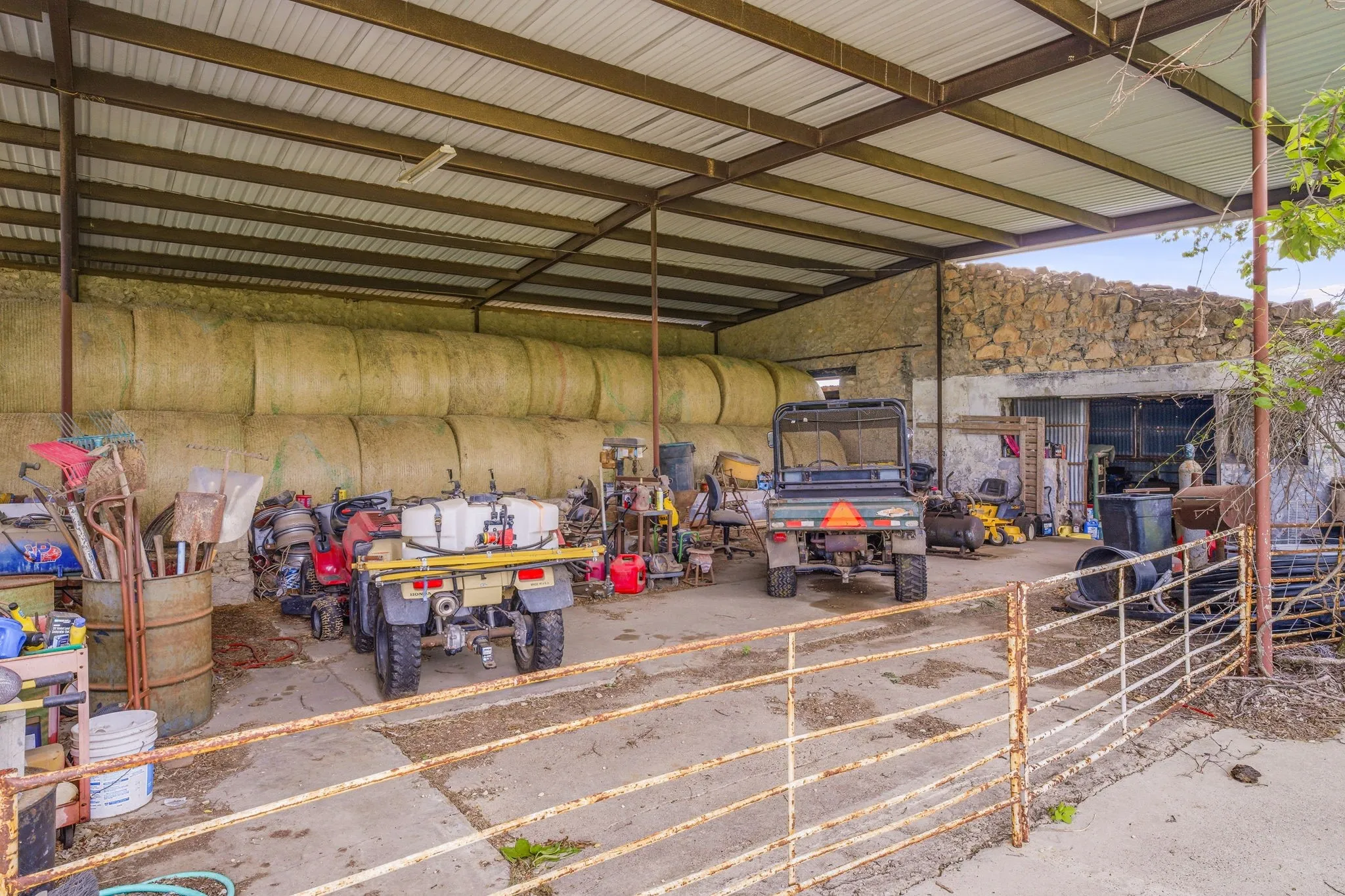 Open-air barn structure featuring a corrugated metal roof supported by metal beams, a concrete slab floor, and a stone wall backdrop