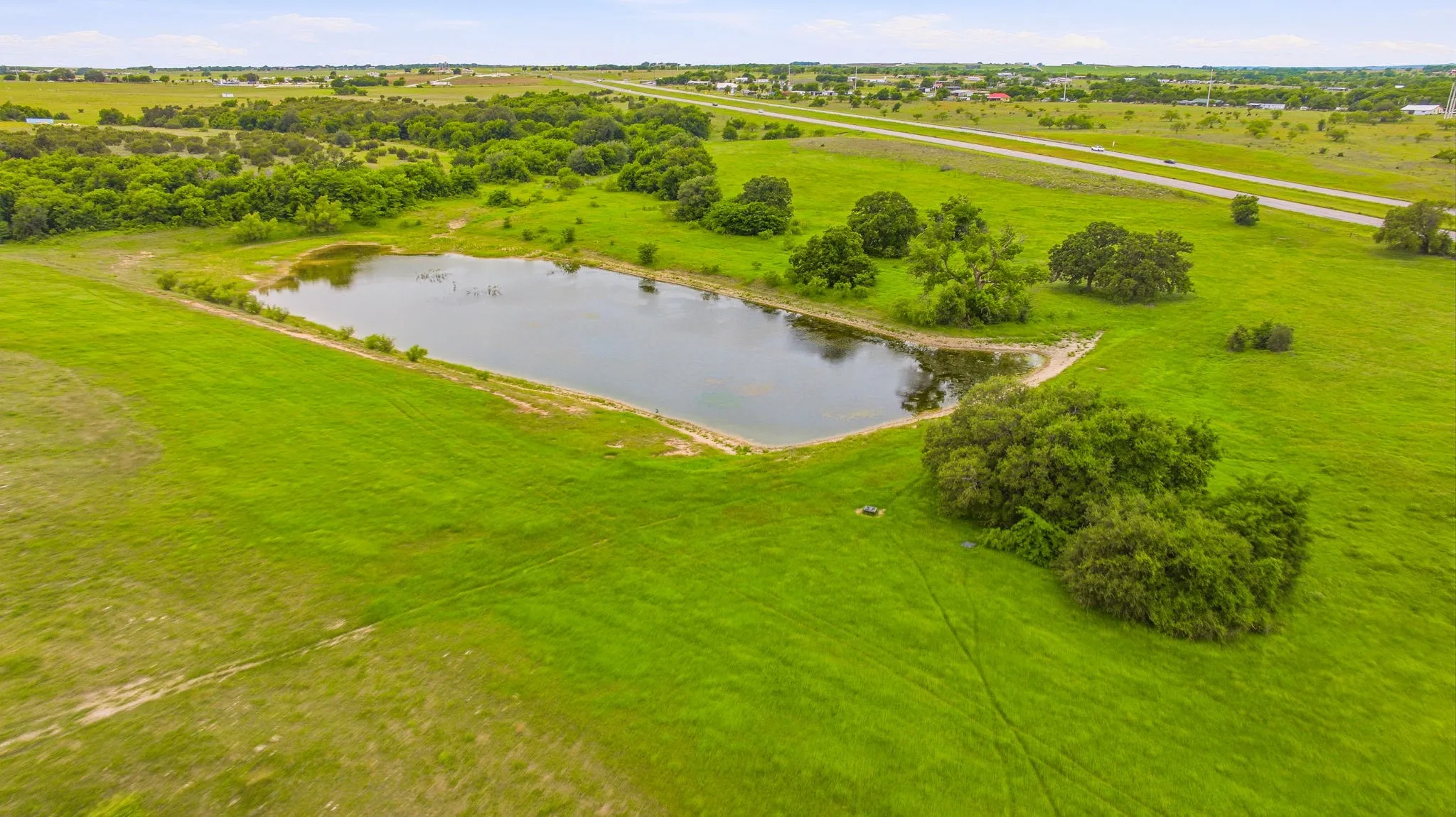 Expansive green landscape featuring a central pond, scattered mature trees, and a distant road