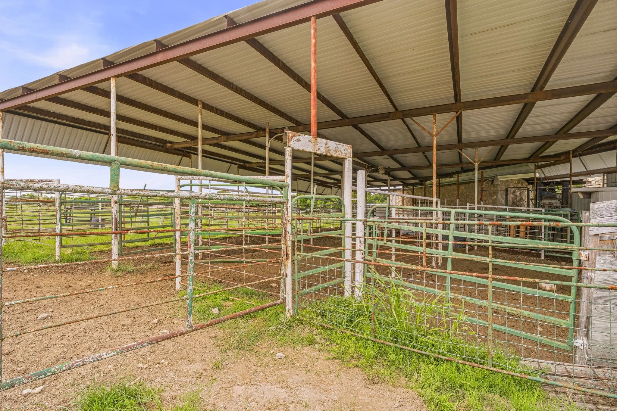 Metal-framed barn structure featuring a corrugated metal roof, multiple interior pens with metal fencing, and a combination of green and rust-colored metal gates