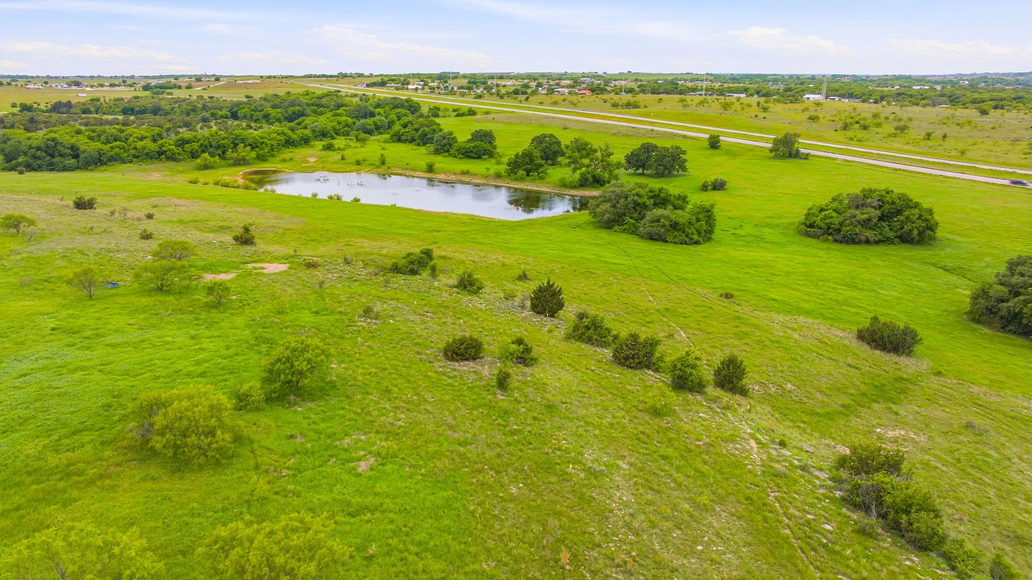 Expansive green landscape featuring a natural pond, mature trees, and open fields with distant roadways