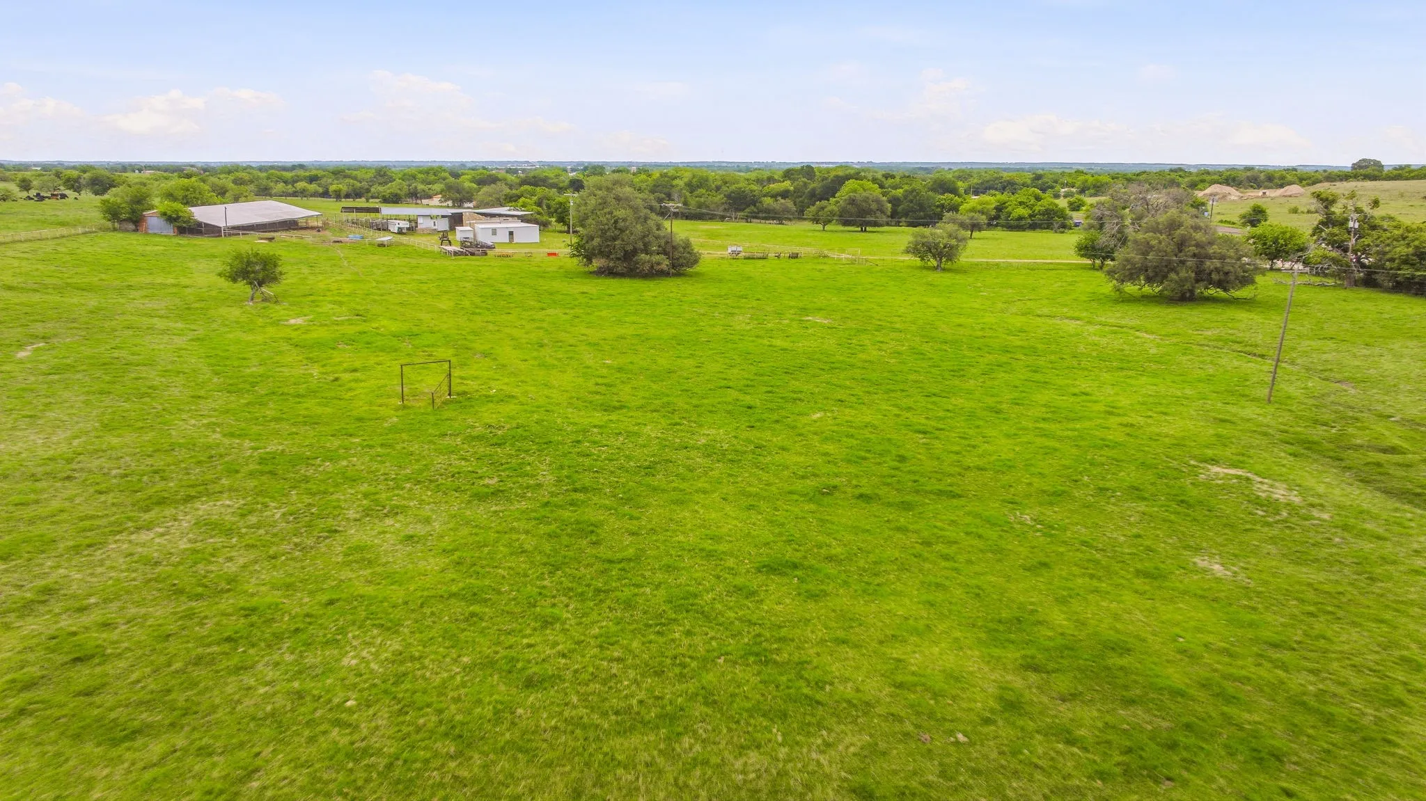 Expansive green acreage featuring a barn structure, secondary outbuildings, mature trees, and open fields under a clear sky