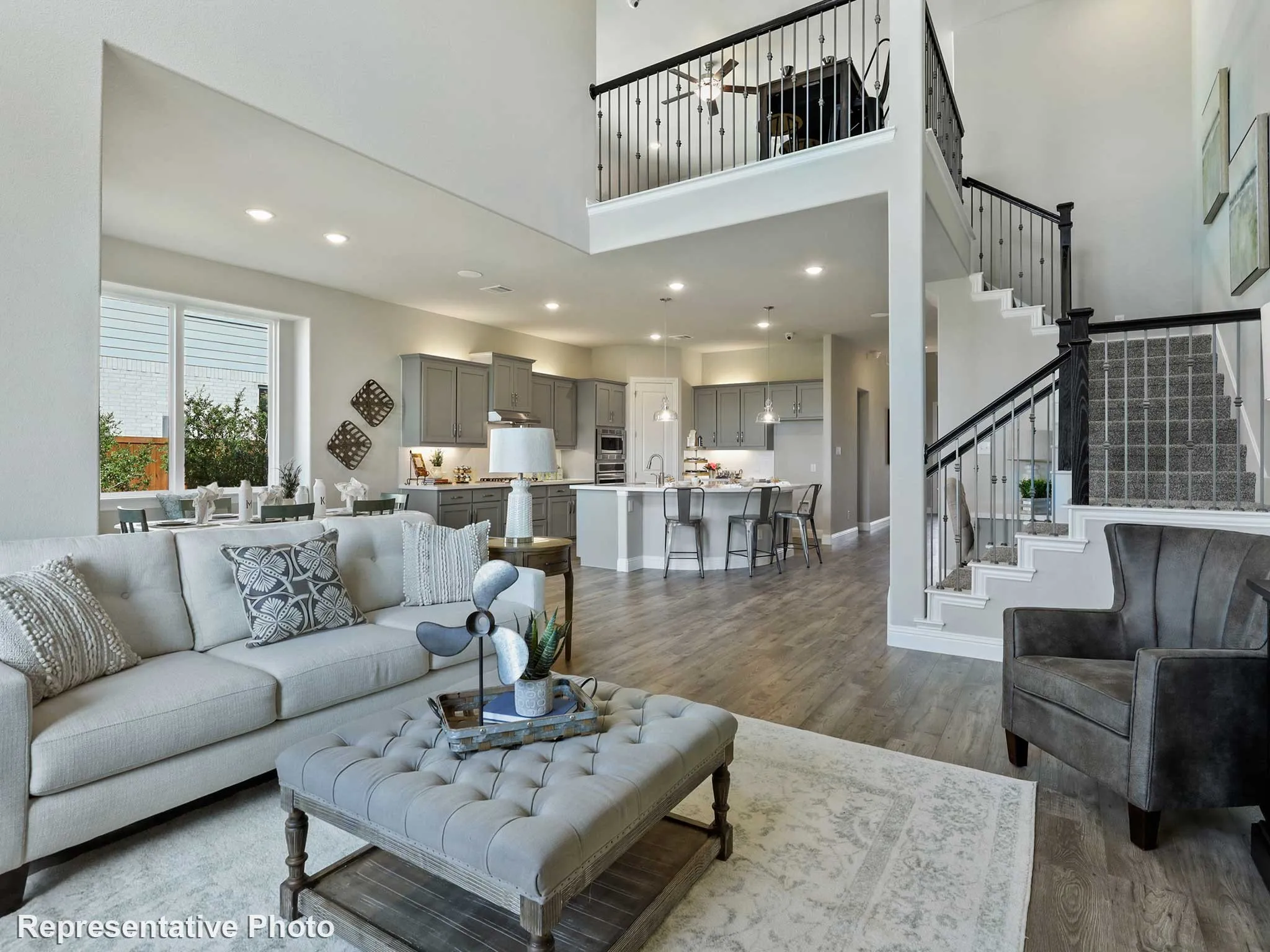 Open-concept living area featuring high ceilings, wood-finish flooring, a two-story staircase with wrought iron balusters, a kitchen with grey cabinetry and an island, and a dining area with ample natural light