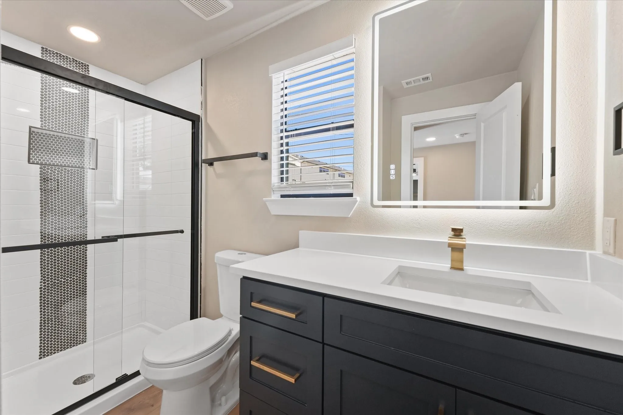Bathroom featuring a dark wood-finish vanity with gold-tone hardware, a white countertop with an integrated rectangular sink, a backlit mirror, and a glass-enclosed shower with black trim and a mosaic tile accent