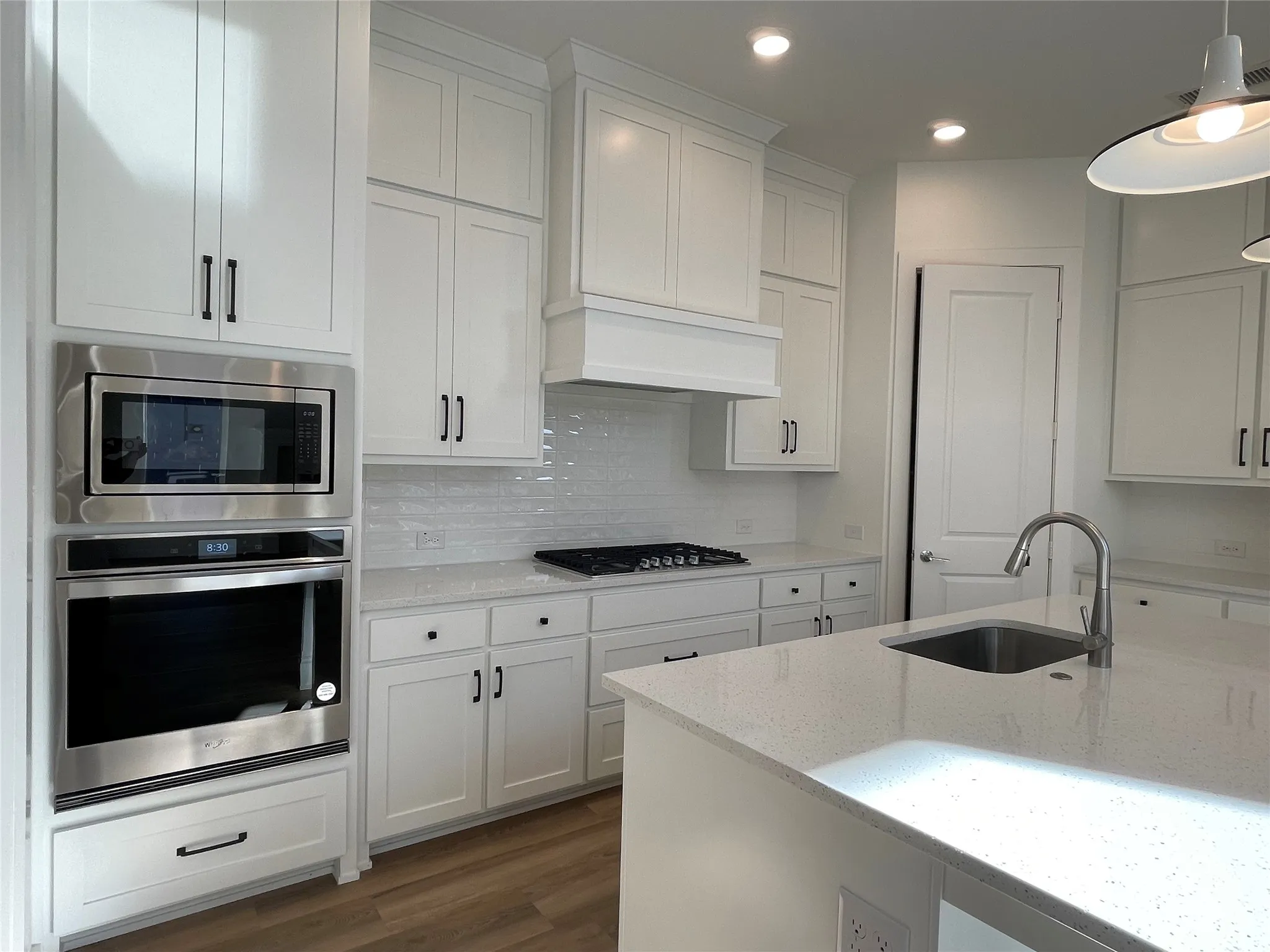Modern kitchen featuring white shaker cabinetry with dark hardware, a stainless steel built-in microwave and oven combination, a gas cooktop, a light-colored speckled countertop, and wood-finish flooring