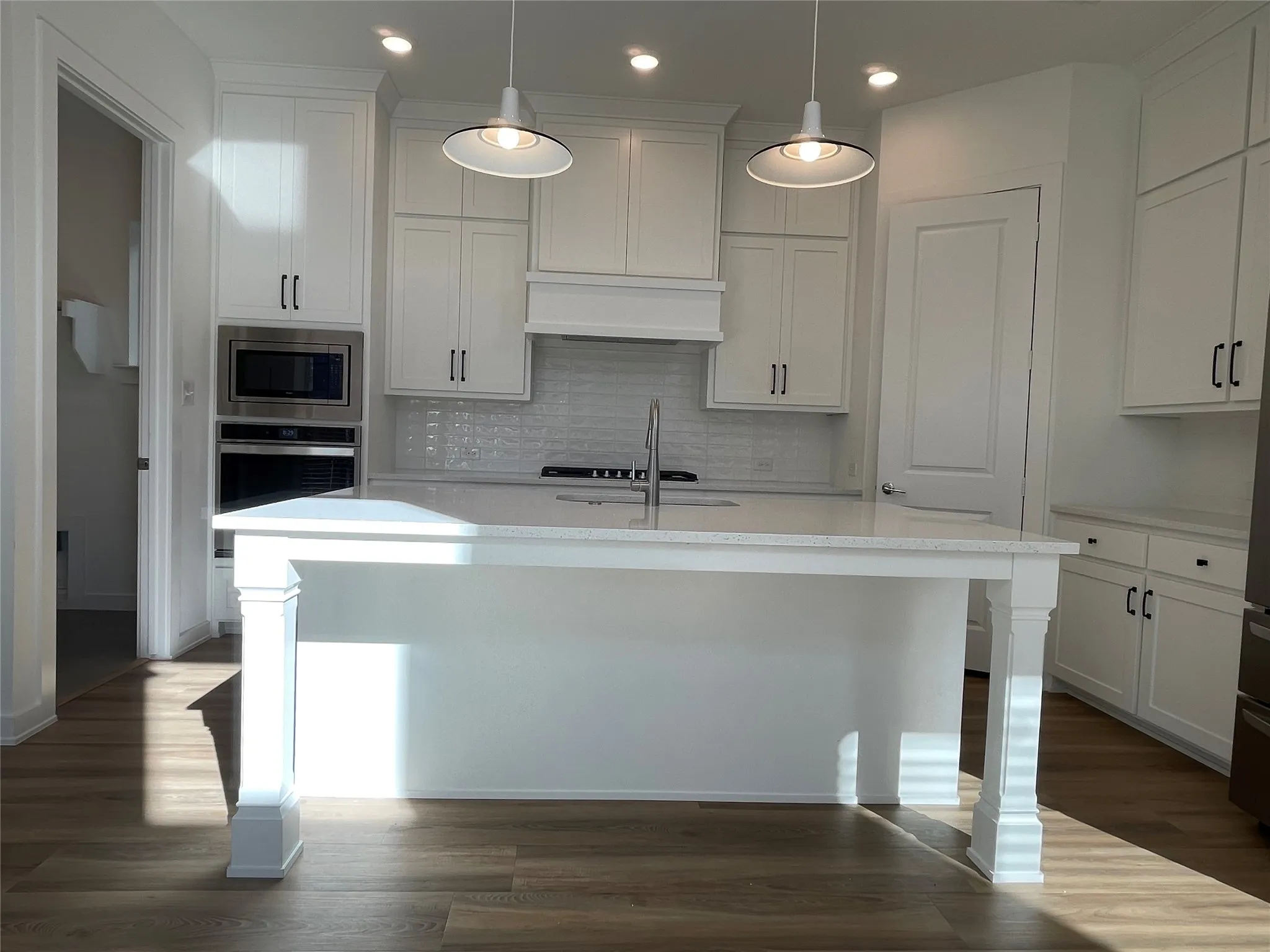 Gourmet kitchen featuring a substantial island with decorative pillar accents, white cabinetry with matte black hardware, stainless steel built-in microwave and oven, a white tile backsplash, and durable wood-finish flooring