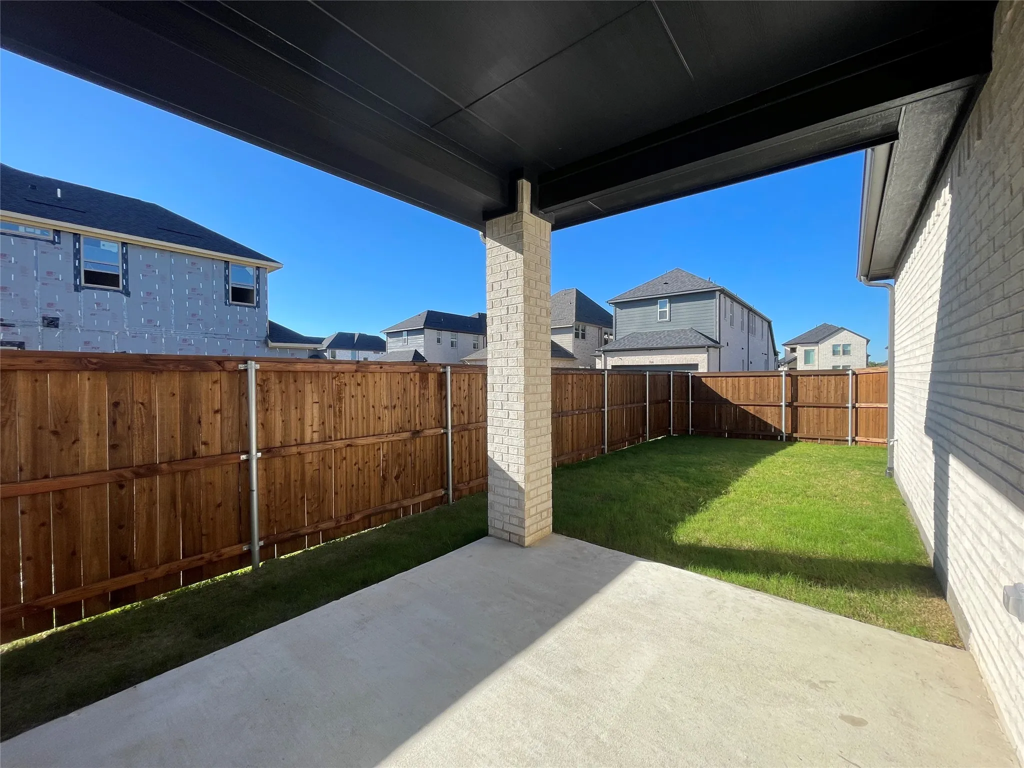 Covered outdoor patio with a concrete surface, bordered by a manicured lawn and a wooden privacy fence