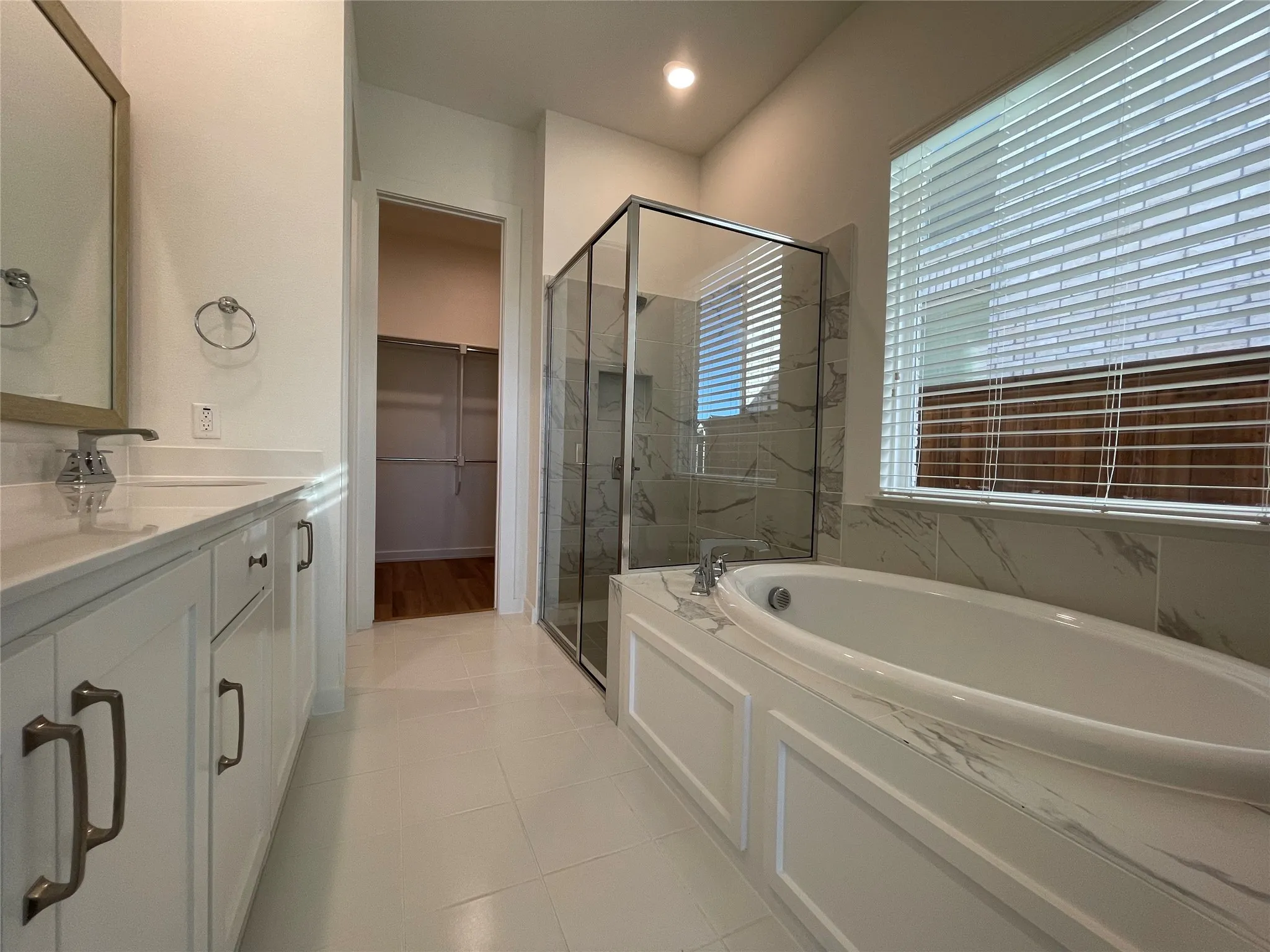 Contemporary bathroom featuring a built-in soaking tub with marble-look surround, a glass-enclosed shower, dual vanities with white cabinetry, and a walk-in closet with wood-finish flooring