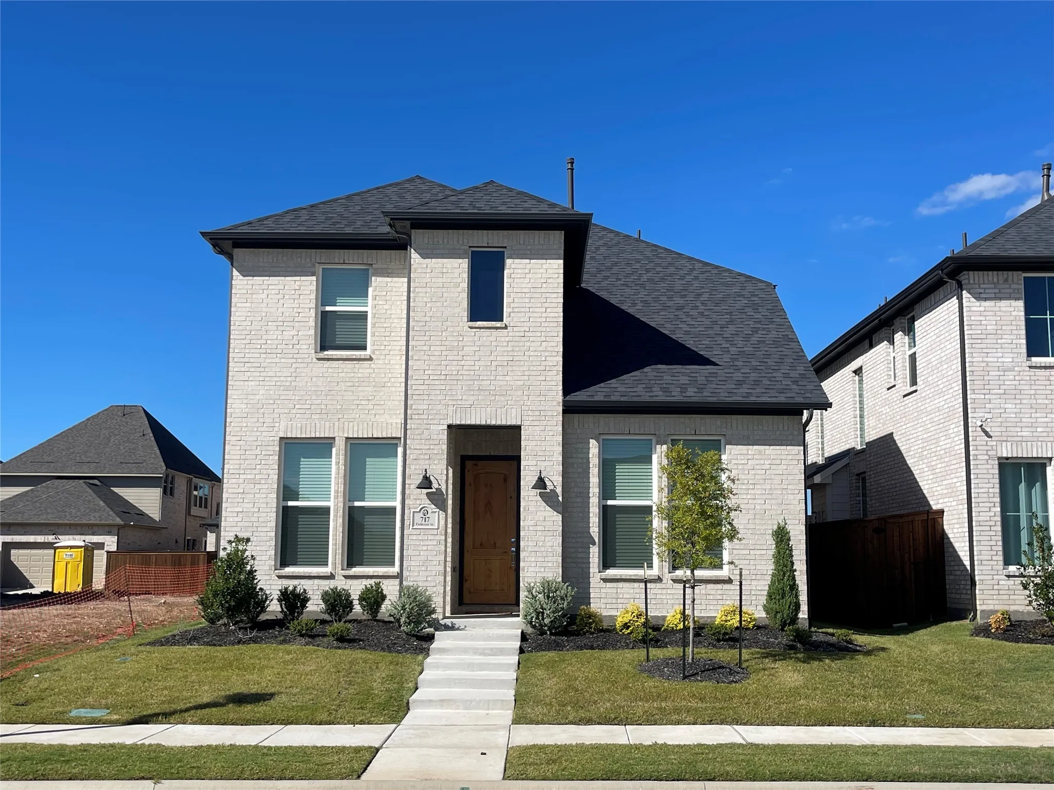 Contemporary two-story residence featuring light-colored brick facade, dark shingle roof, dark wood entry door, and manicured front landscaping