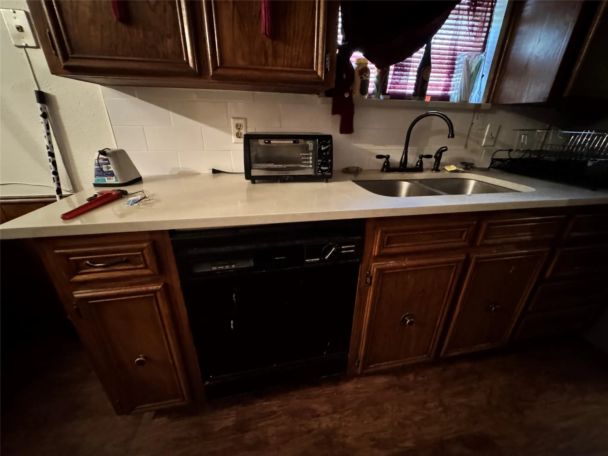 Kitchen featuring dark wood-finish cabinetry, light-toned countertops, a tile backsplash, a double basin sink with a dark-finish faucet, and a dishwasher
