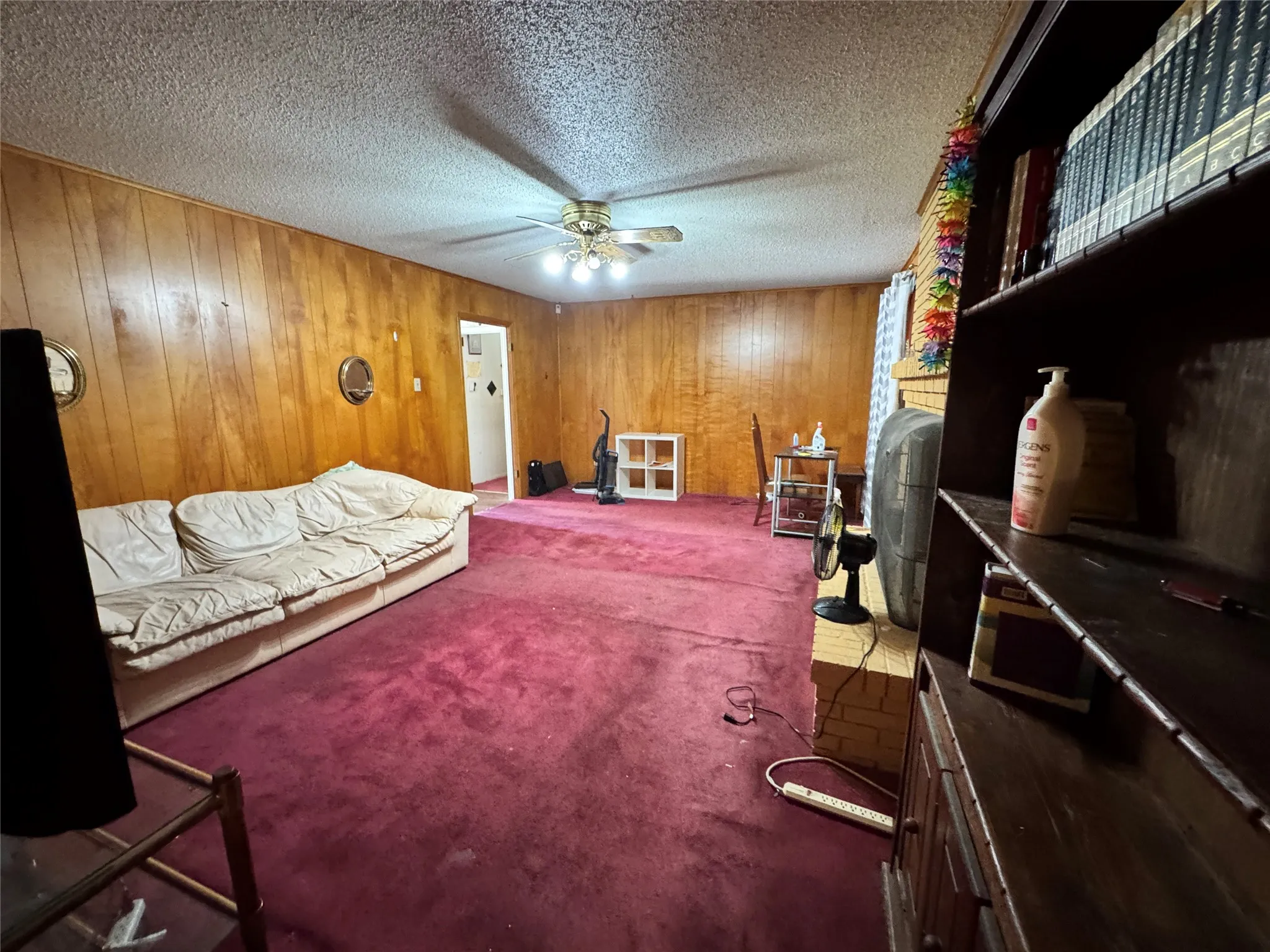 Living area featuring wood paneling, a brick fireplace, and a ceiling fan with lighting and red carpeting