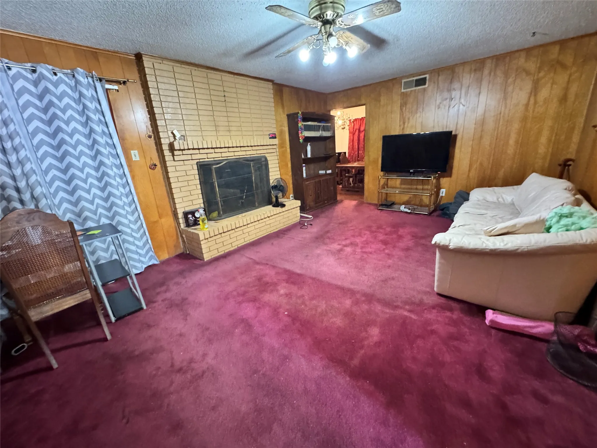 Living area featuring wood paneling, a brick fireplace, and a ceiling fan with lighting and red carpeting