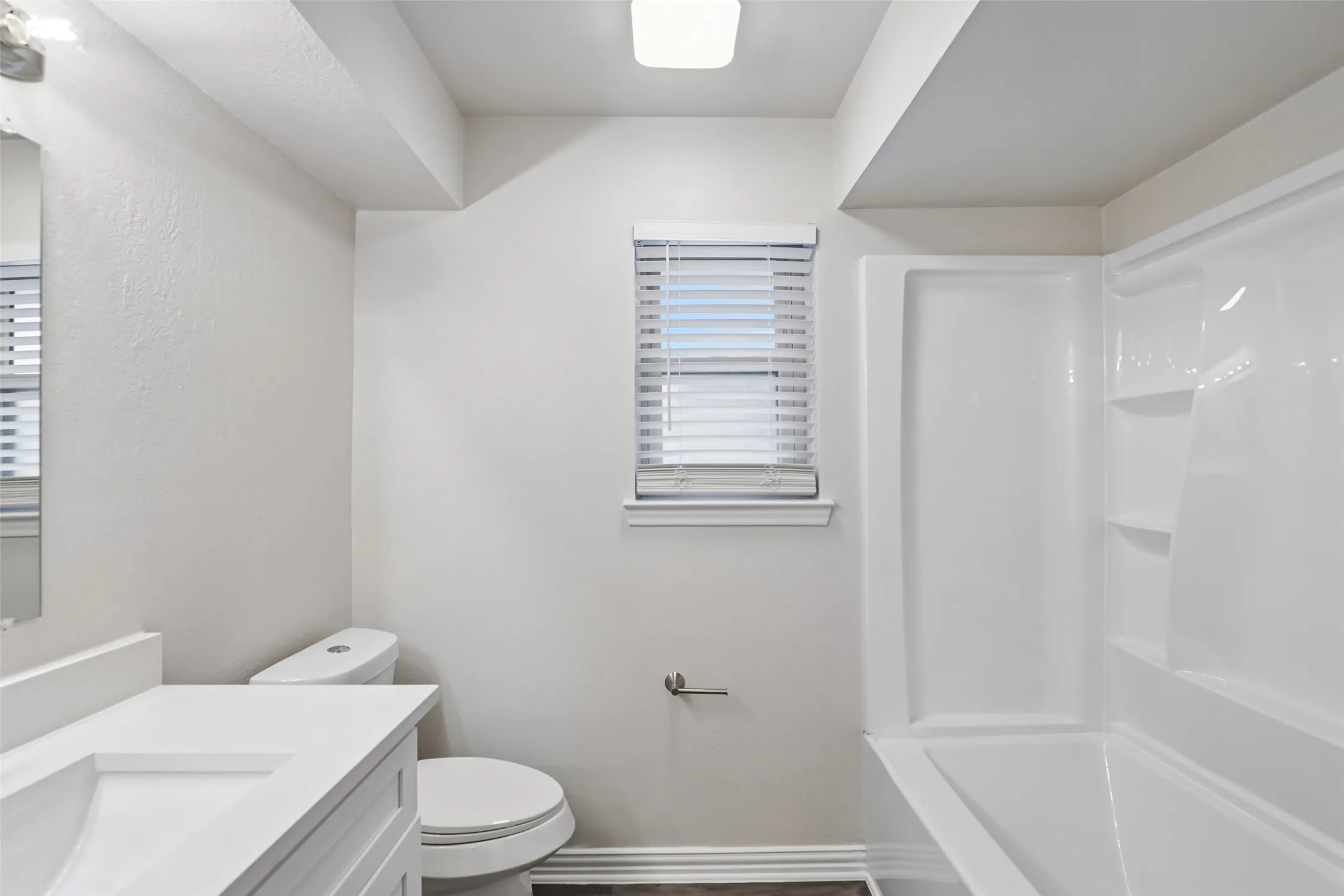 Bathroom featuring a white integrated sink vanity, a toilet, a window with blinds, a white tub, and wood-finish flooring
