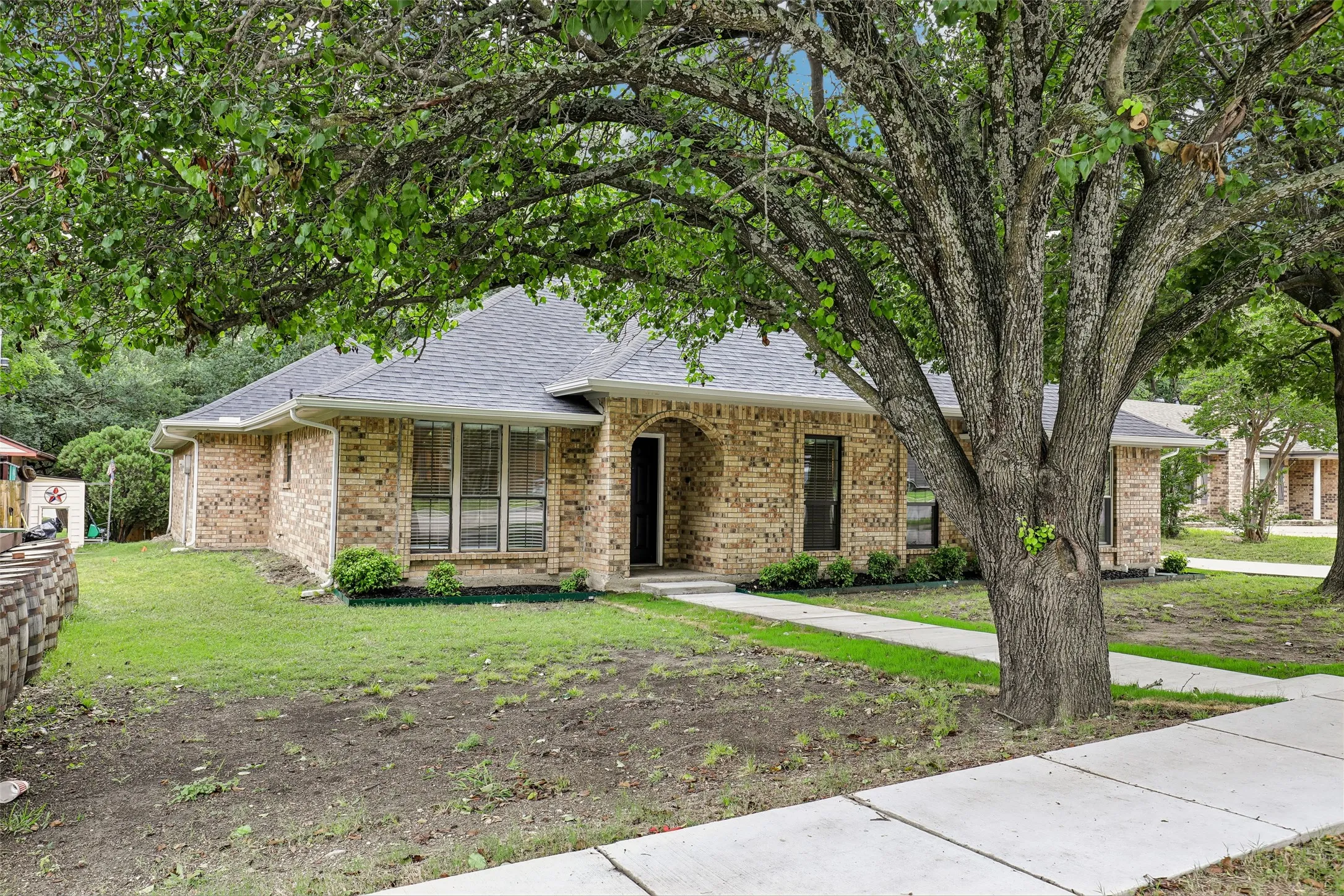 Brick exterior with a dark shingle roof, featuring mature trees providing ample shade