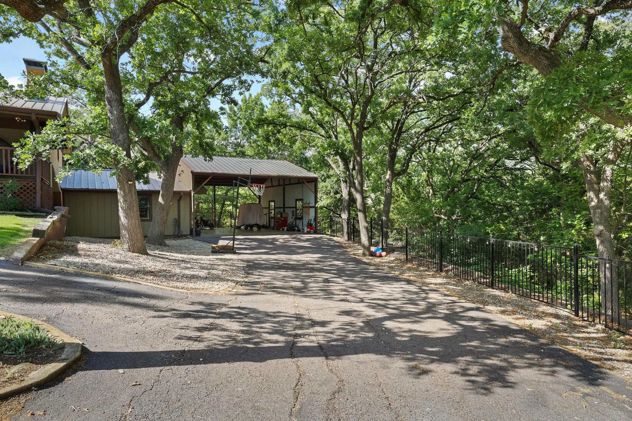 Paved driveway leading to a covered carport, flanked by mature trees and a black metal fence