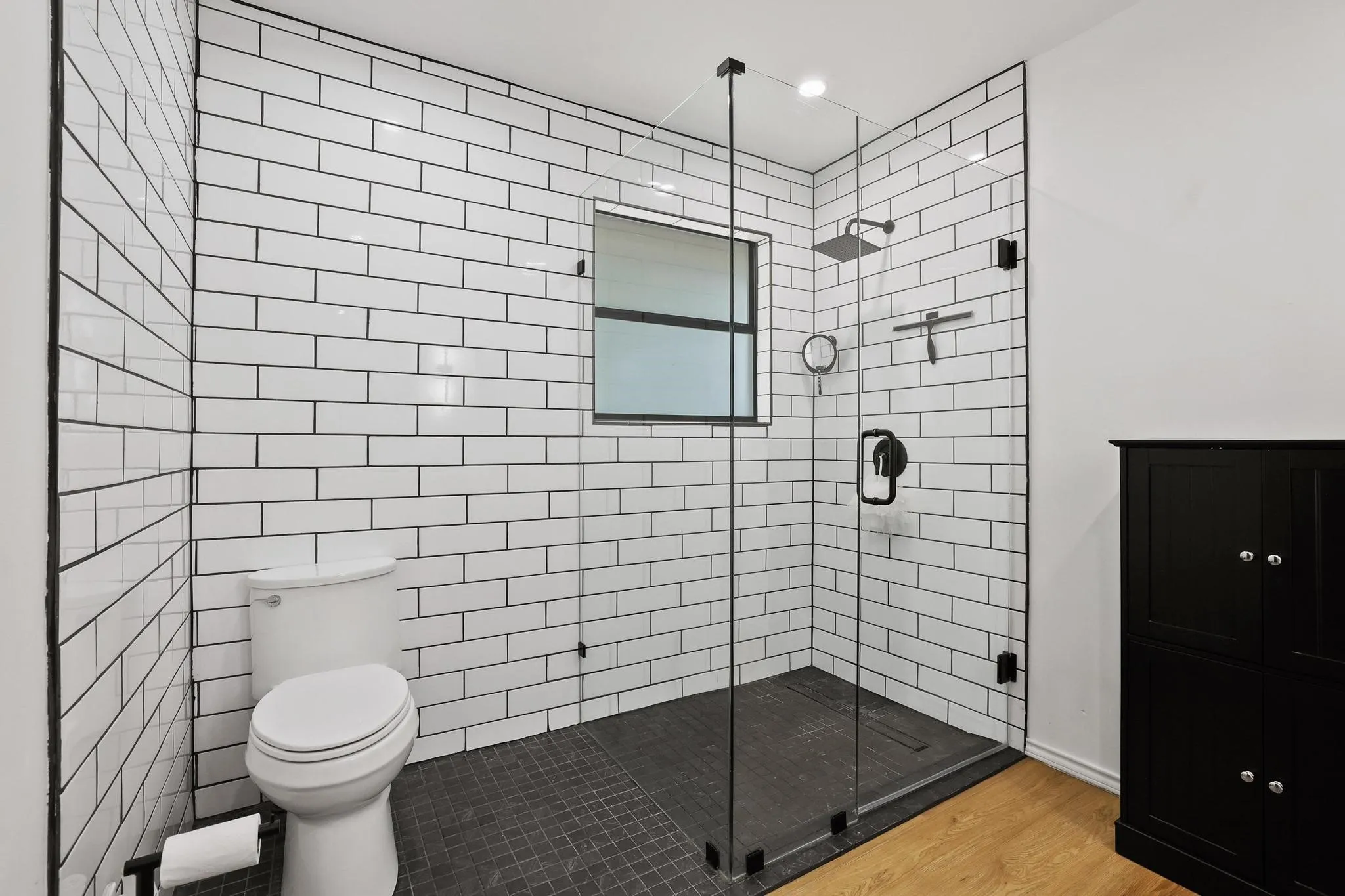 Spacious bathroom featuring a glass-enclosed shower with matte black fixtures, white subway tile walls with dark grout, dark tile shower flooring, and a window for natural light