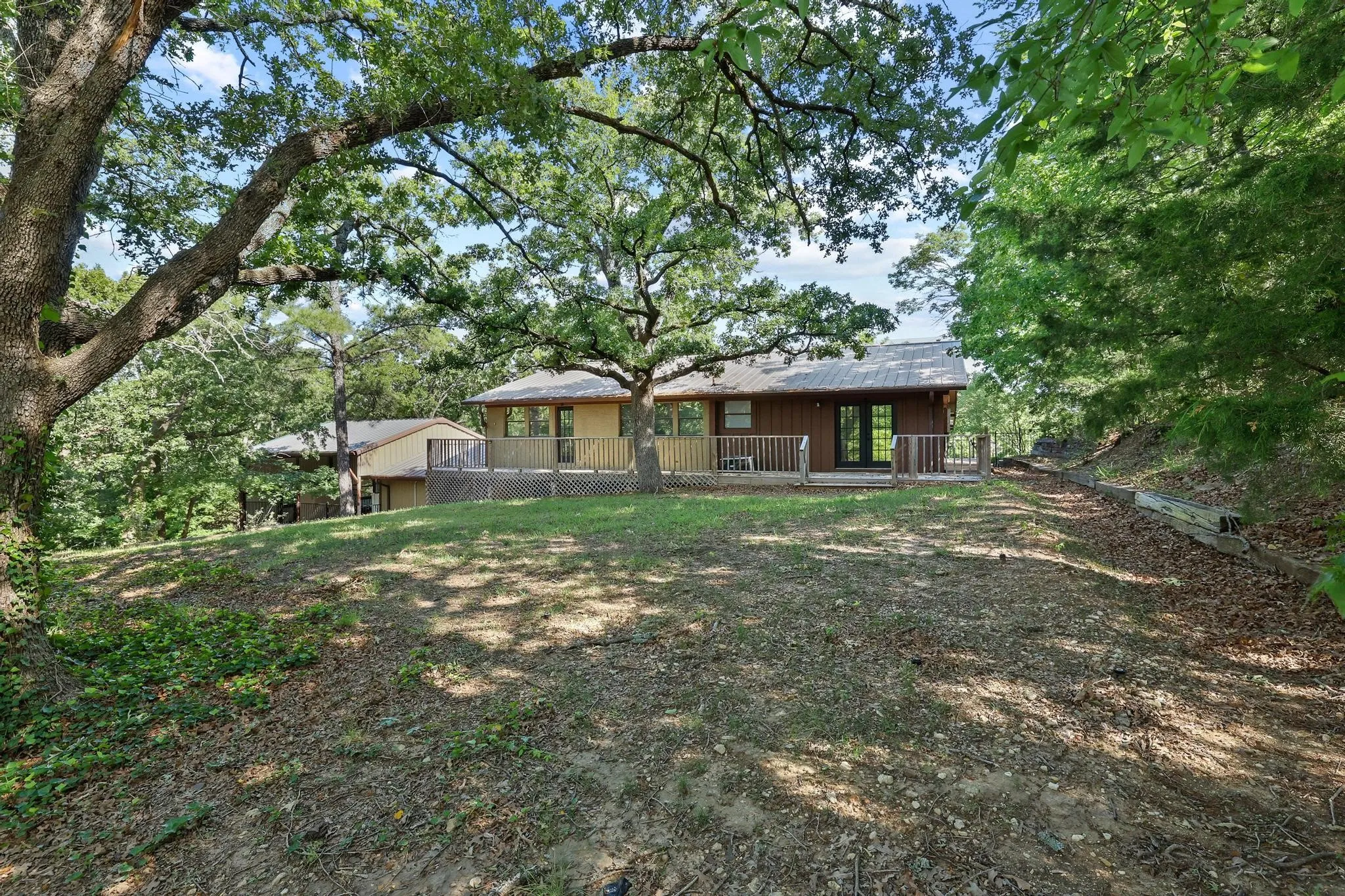 Elevated property featuring a wooden deck, mature trees, and a sloped landscape