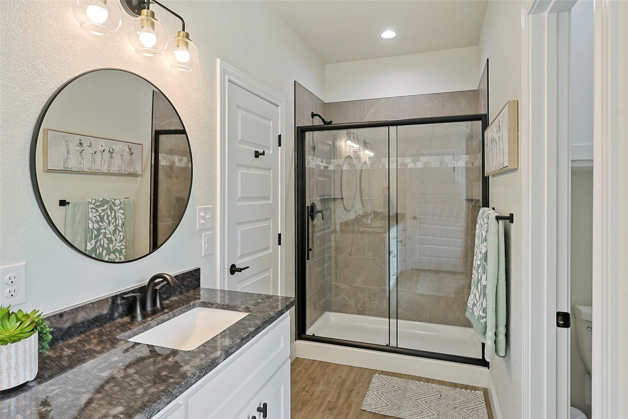 Bathroom featuring a vanity with a dark stone countertop and undermount sink, a circular mirror, and a sliding glass shower enclosure with tiled walls
