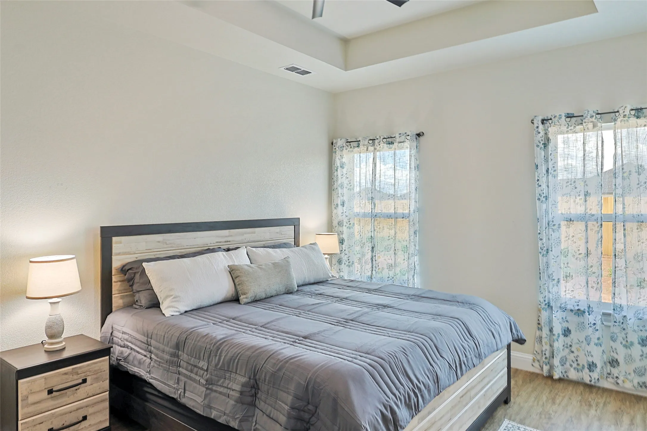 Spacious room featuring wood-finish flooring, a tray ceiling, and two large windows with sheer curtains