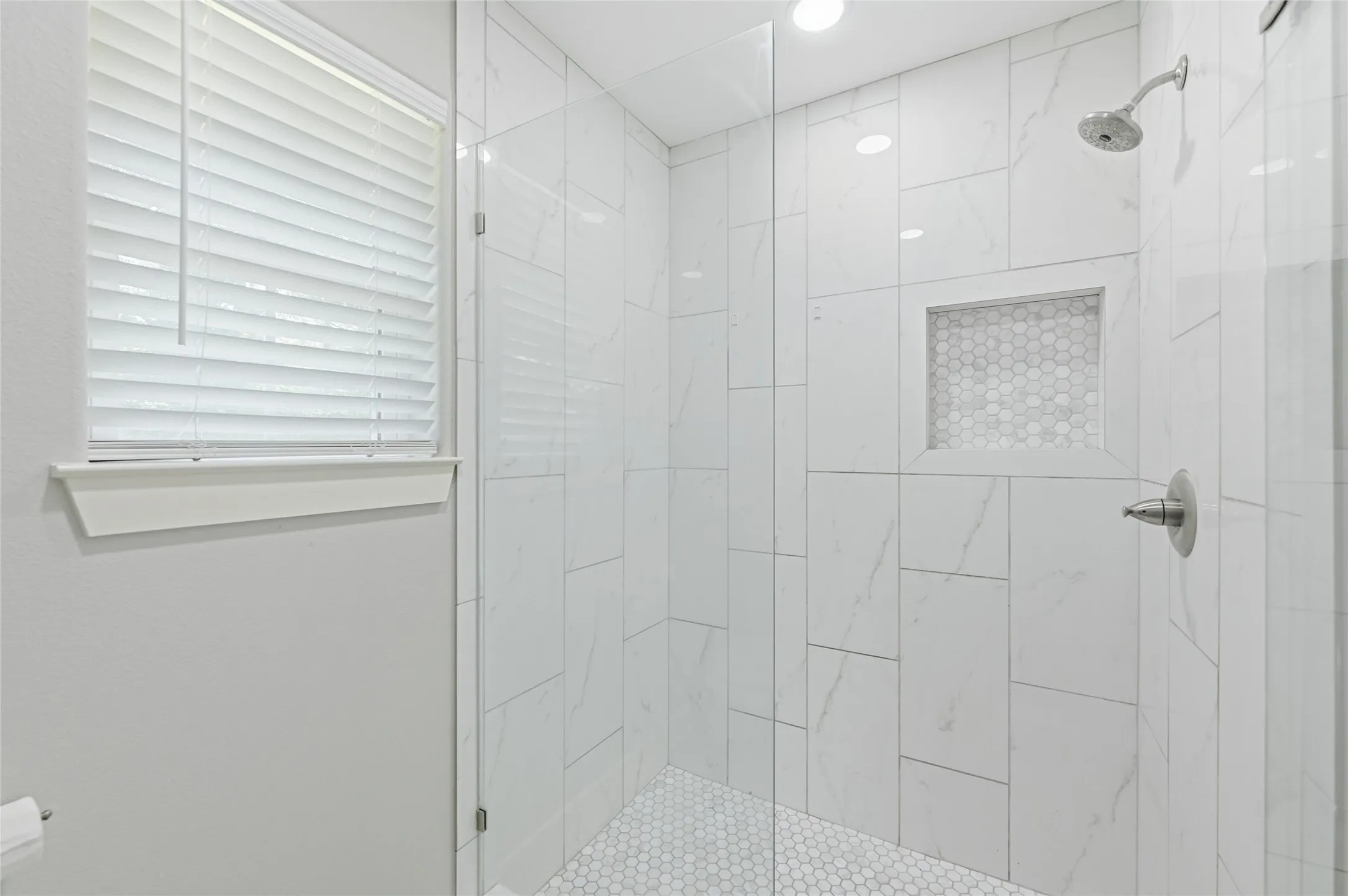 Walk-in shower featuring a frameless glass enclosure, large format white tile walls, a hexagon tile shower niche, and hexagon tile flooring