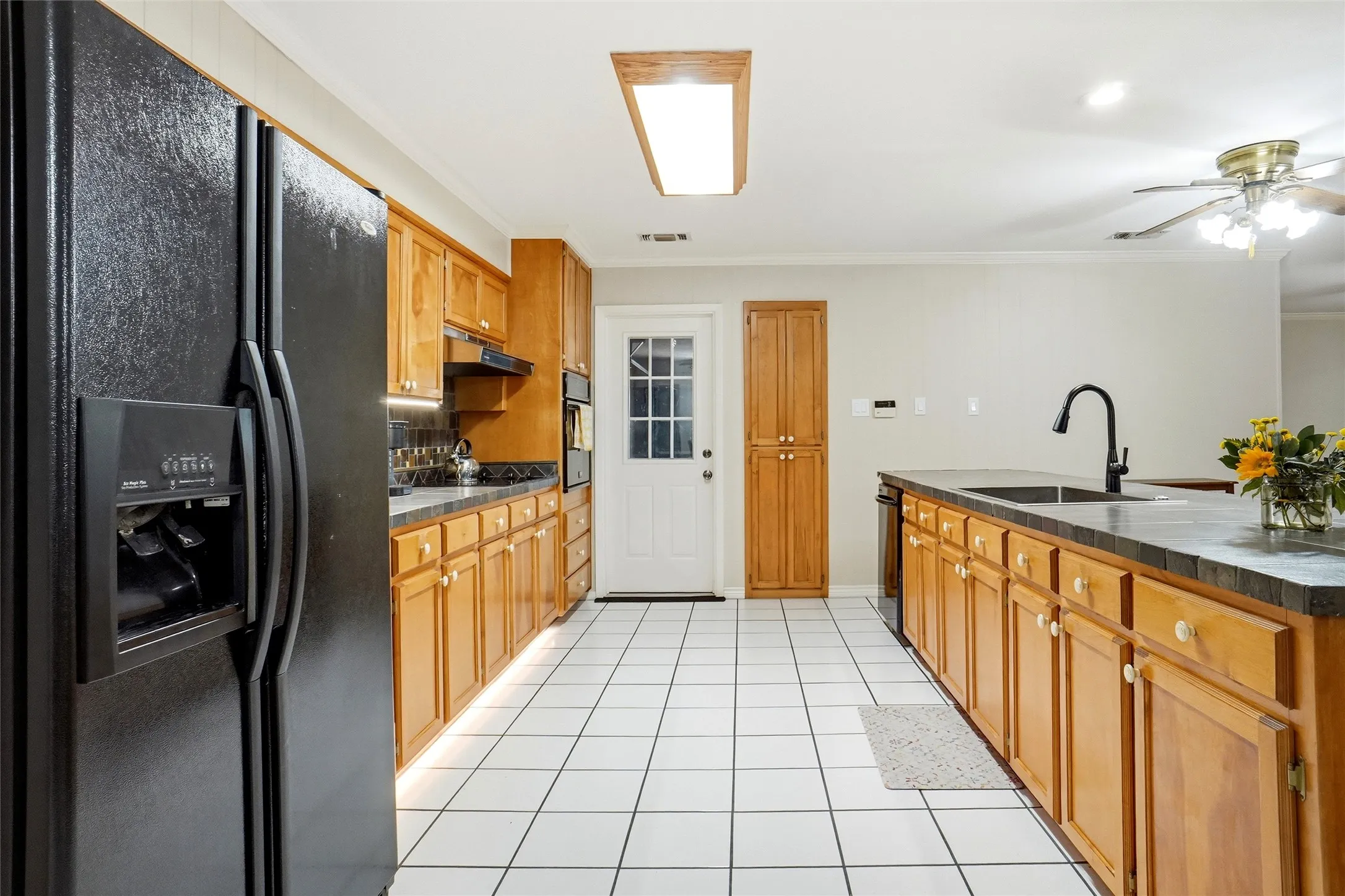 Kitchen featuring white square tile flooring, wood cabinetry with light hardware, black countertops, a black side-by-side refrigerator, and a built-in oven