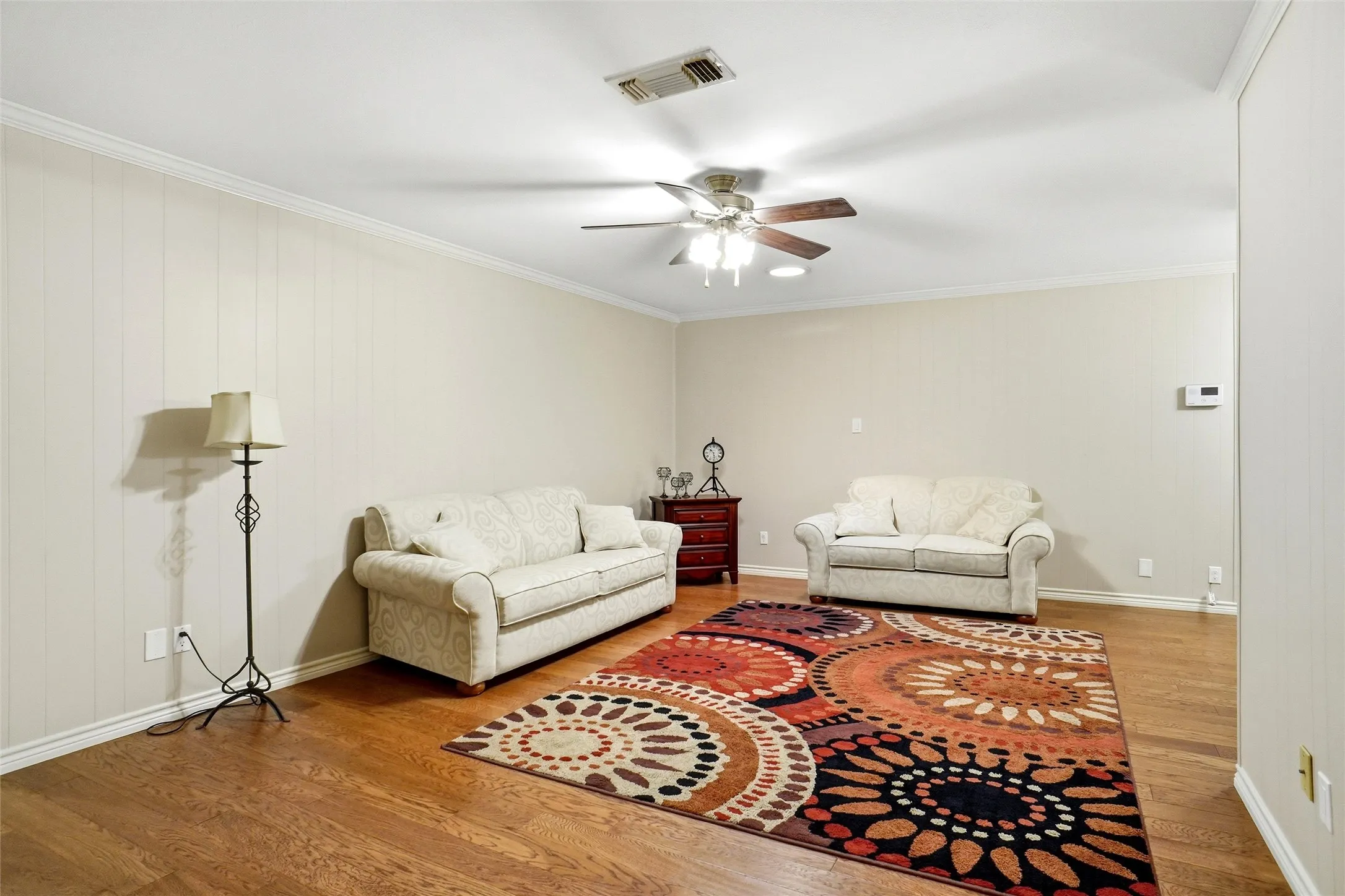 Spacious room featuring wood-finish flooring, a ceiling fan with light fixture, vertical paneling on one wall, and crown molding