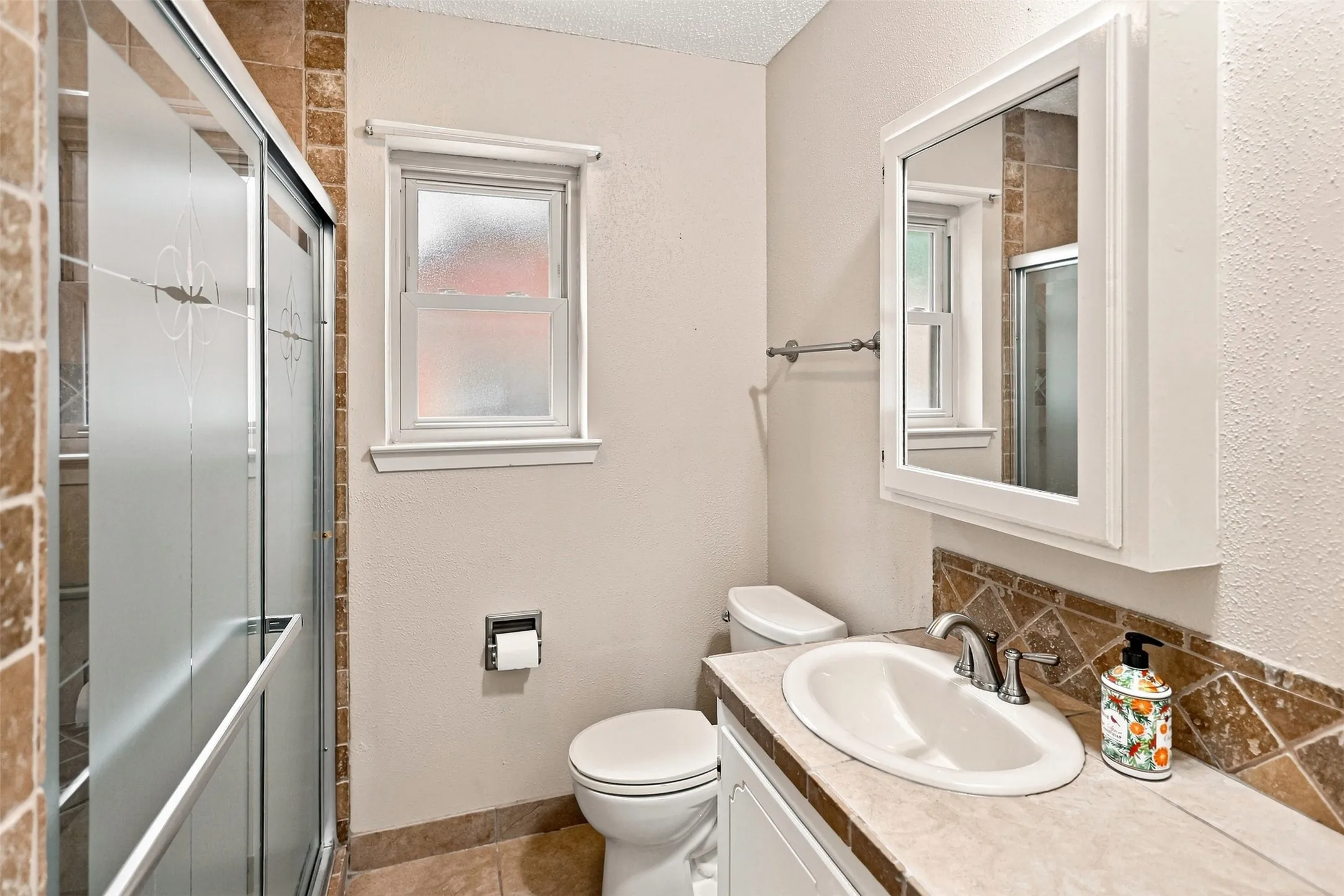Hall Bath shower with frosted glass enclosure, single-pane window with white trim, wall-mounted medicine cabinet with mirror, vanity with an integrated sink, and a tiled backsplash