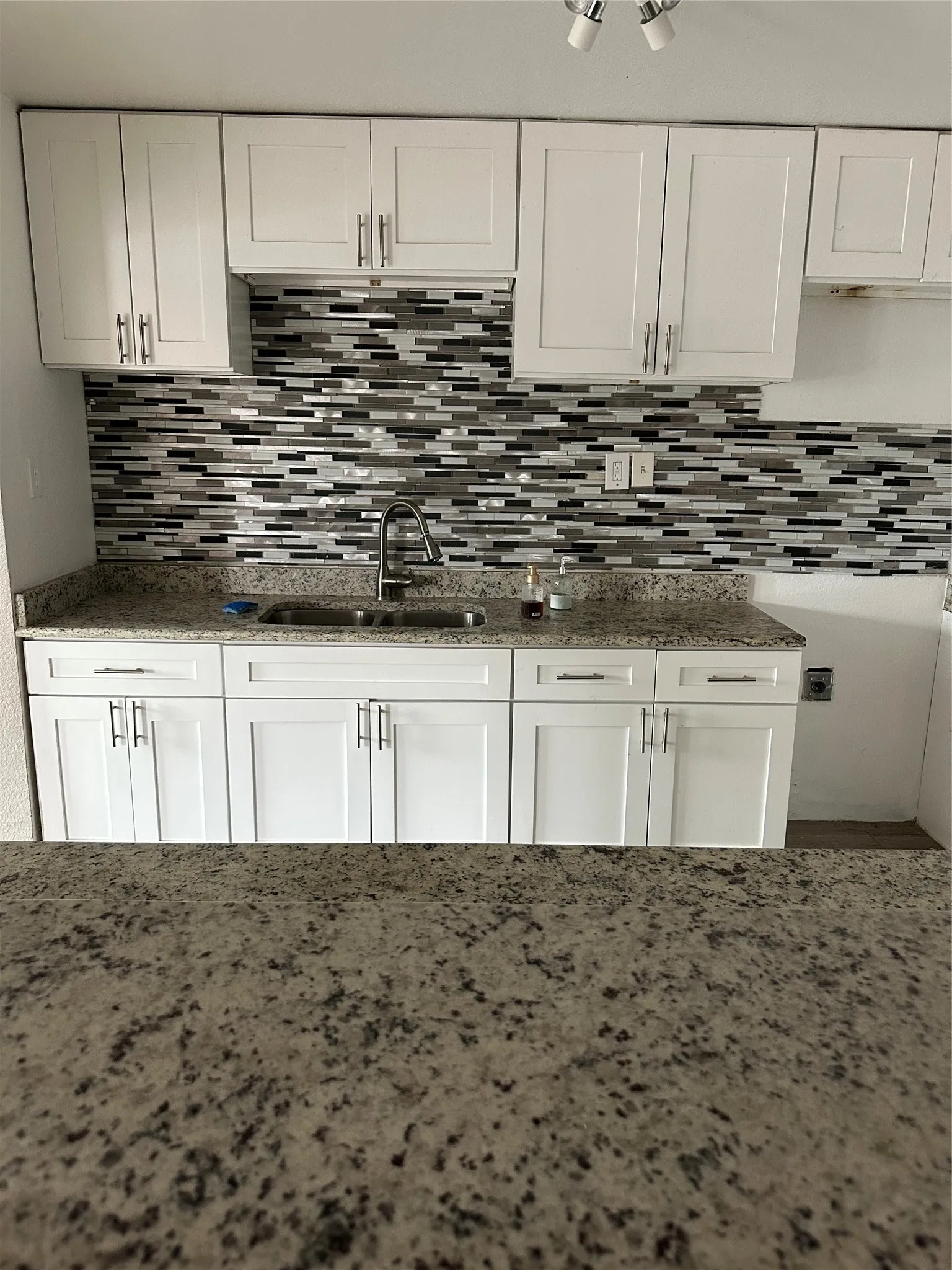 Kitchen featuring white shaker-style cabinetry with brushed nickel hardware, speckled granite countertops, a mosaic tile backsplash in varying shades, and a stainless steel double basin sink with a gooseneck faucet