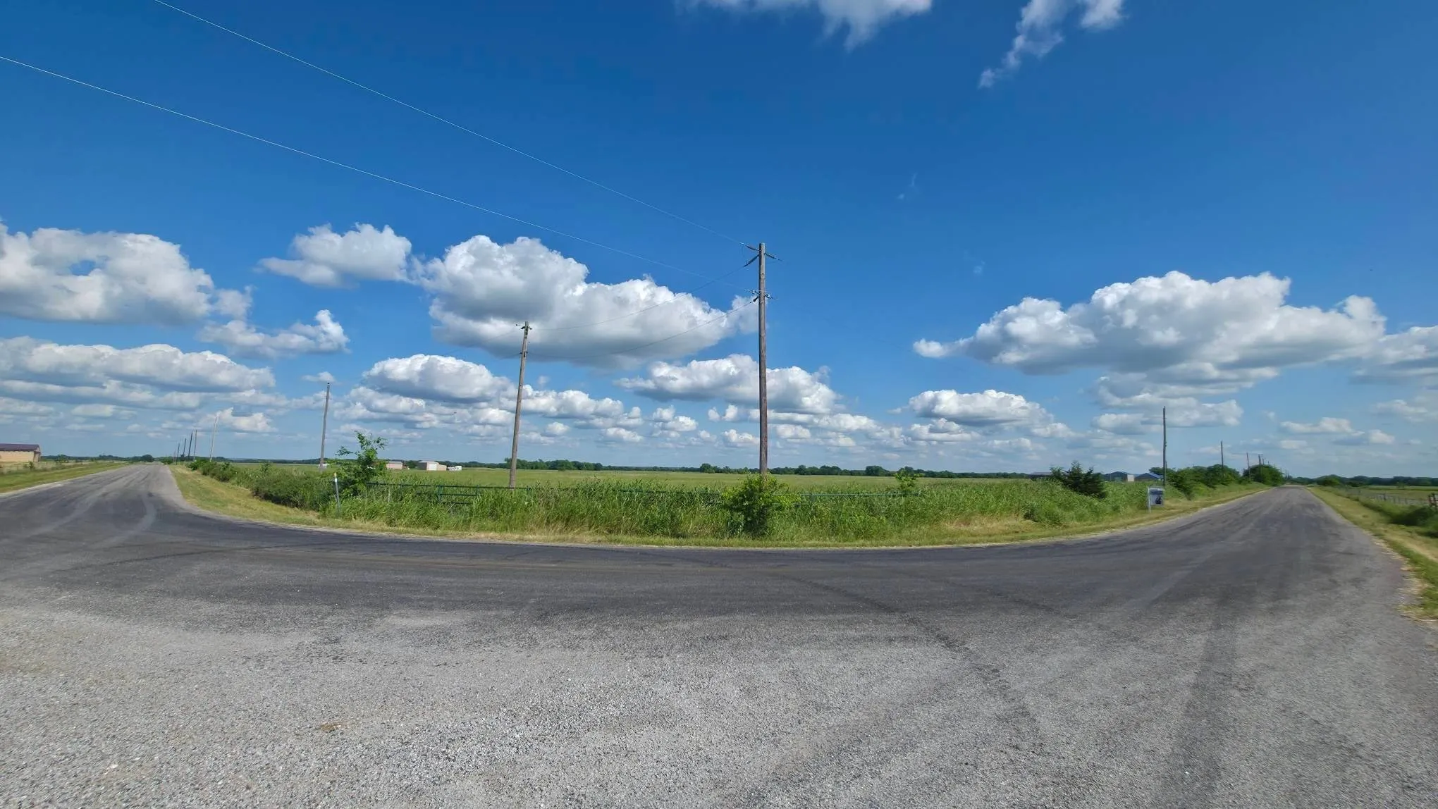 Rural intersection featuring an asphalt road with a gravel shoulder, expansive green fields, and overhead utility lines