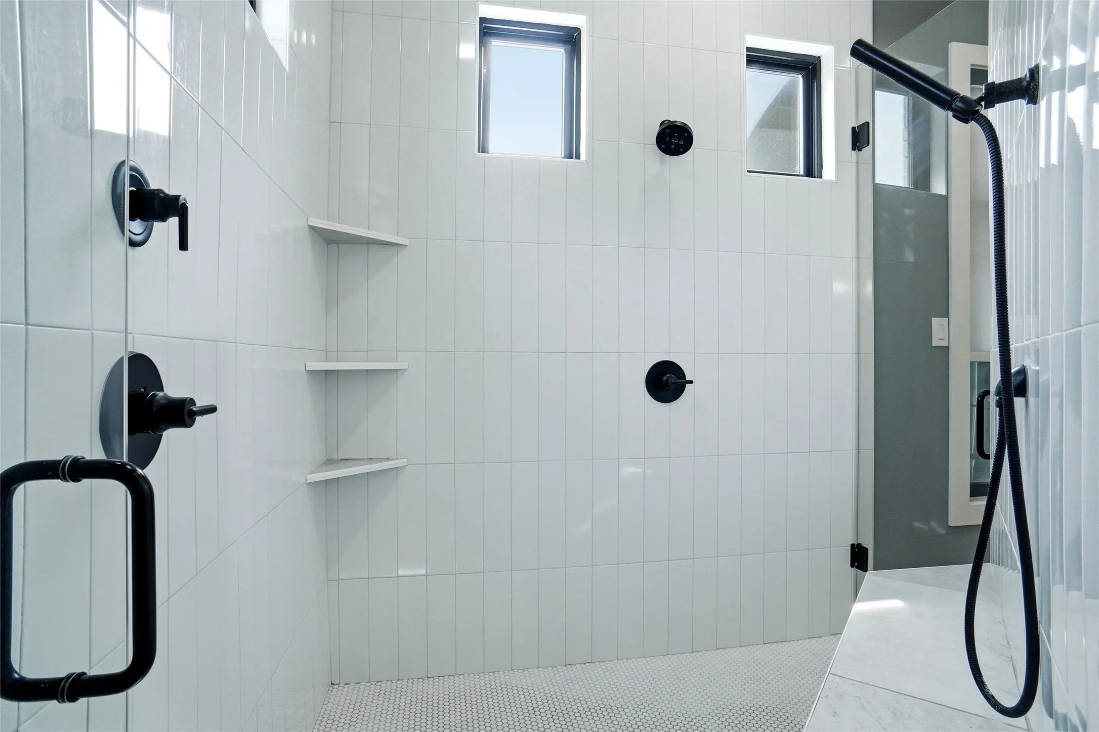 Primary Bath Shower enclosure featuring full-height white tile walls, matte black fixtures, built-in corner shelving, a handheld shower fixture, and a mosaic tile floor