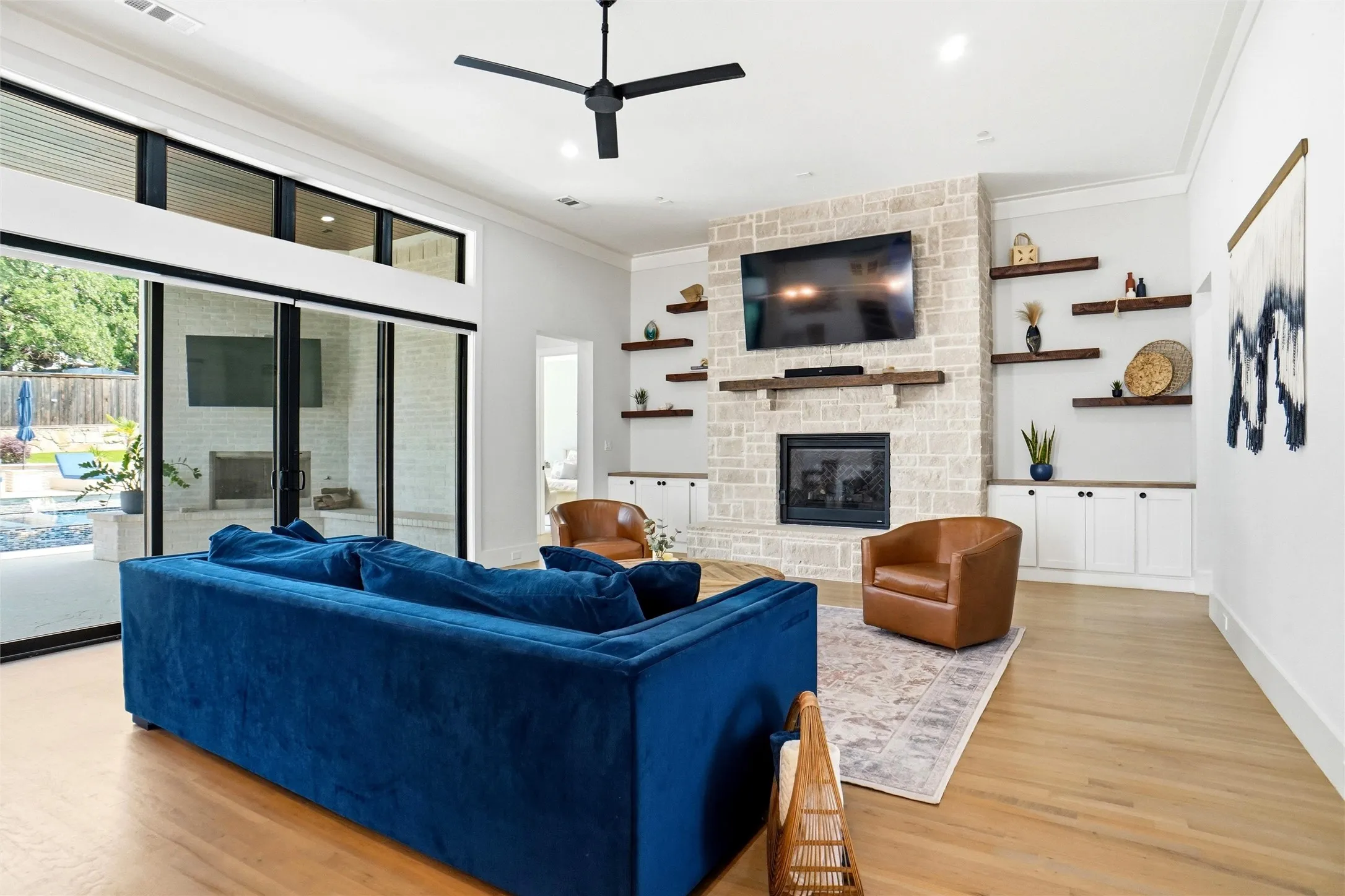 Spacious living area featuring a stone-clad fireplace, built-in shelving and cabinetry, wood-finish flooring, a contemporary ceiling fan, and large sliding glass doors opening to the backyard