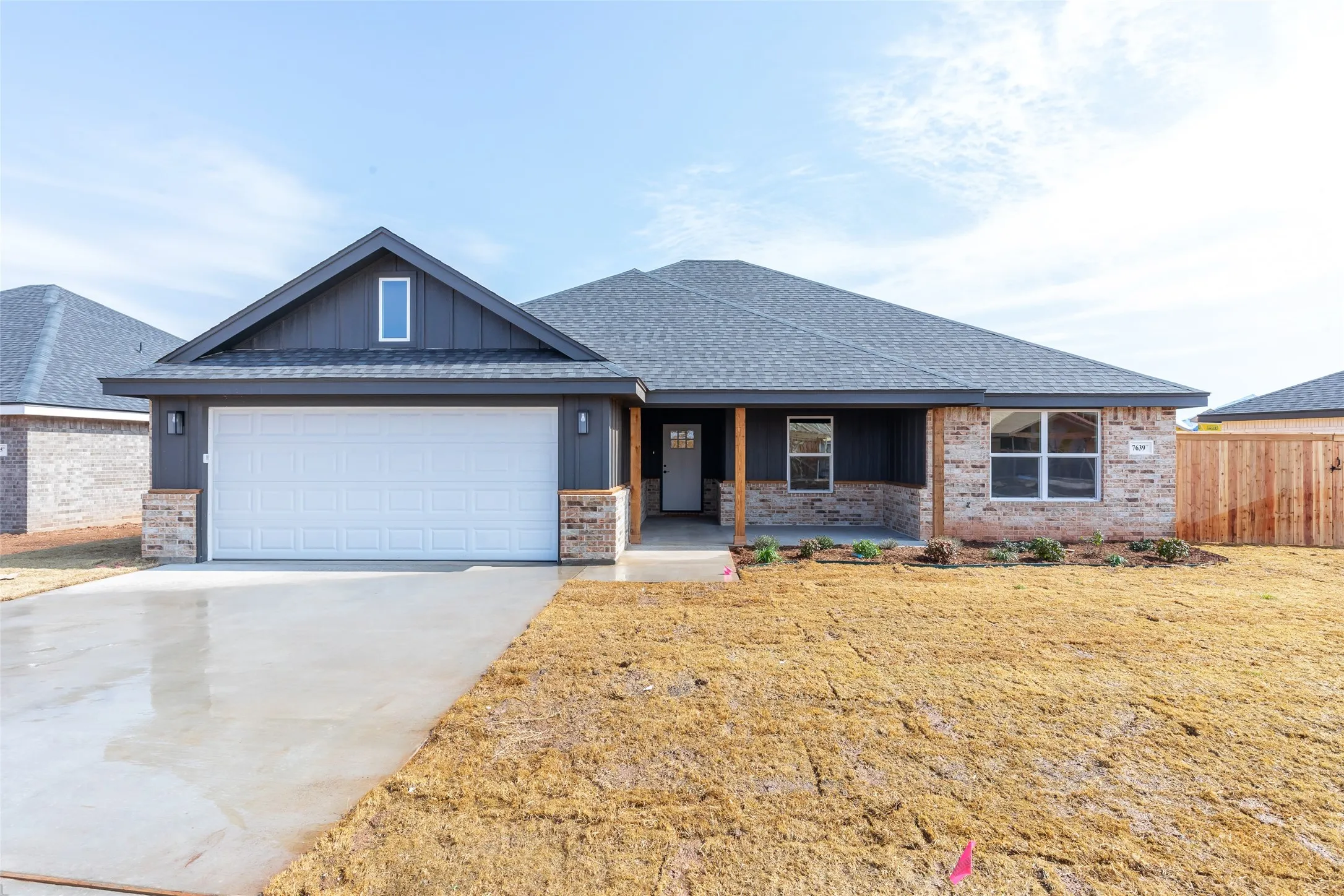 Modern ranch-style home featuring a two-car garage, dark gray siding, brick accents, a covered front porch, and a privacy fence