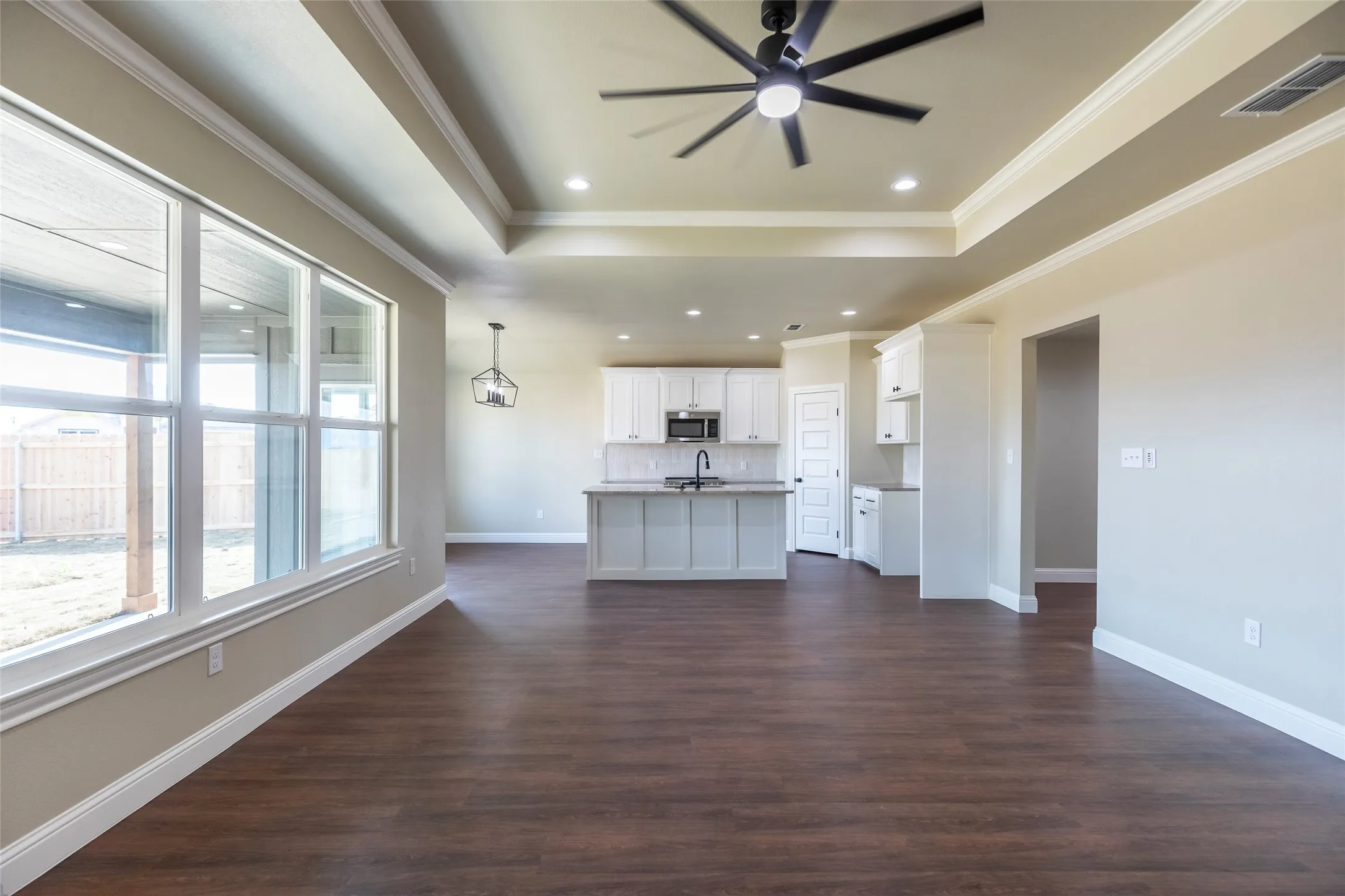 Expansive open-concept living area featuring wood-finish flooring, a tray ceiling with recessed lighting and a ceiling fan, and large windows