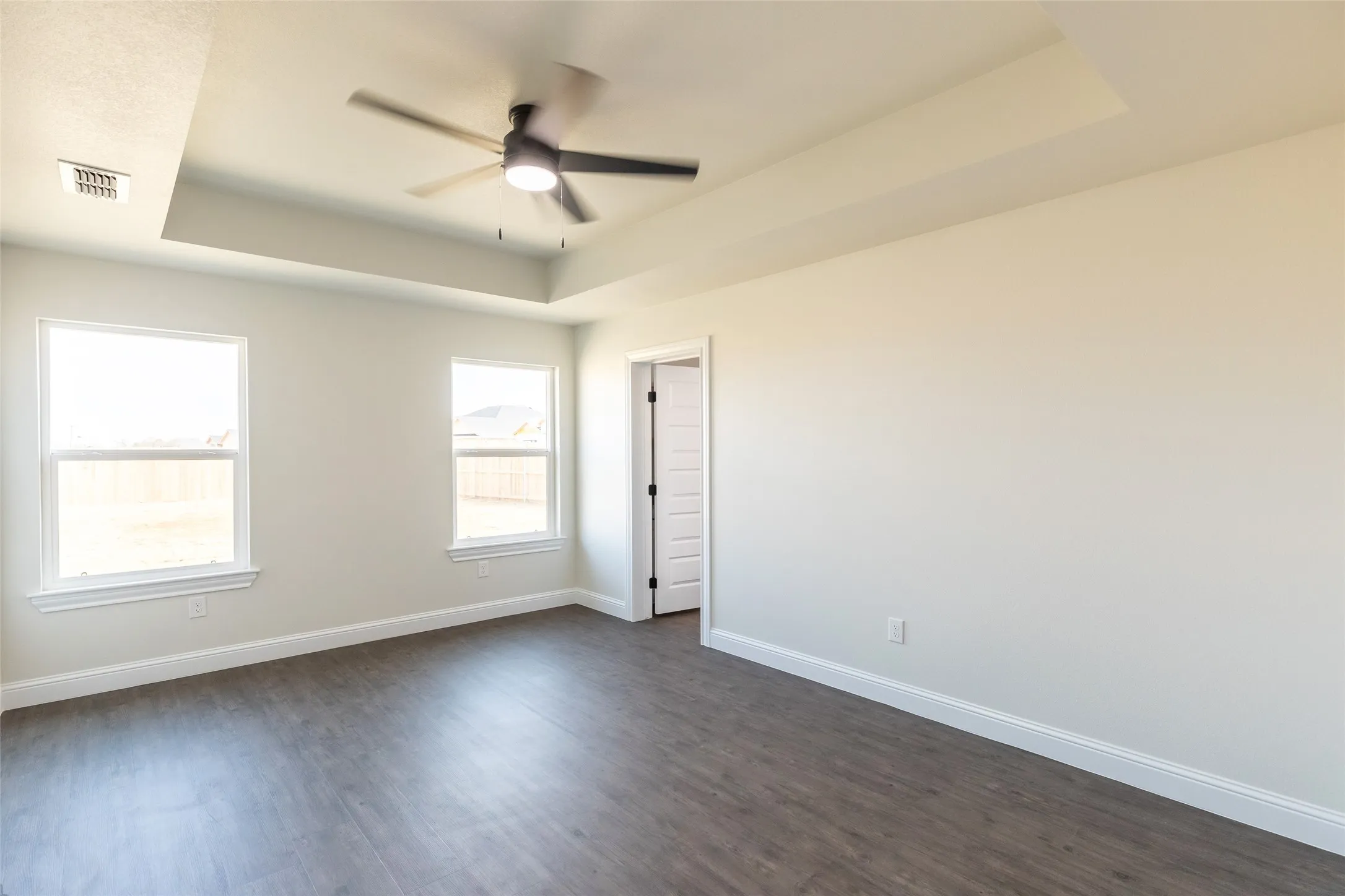 Spacious room featuring tray ceiling with a modern ceiling fan, wood-finish flooring, two large windows, and a panel door
