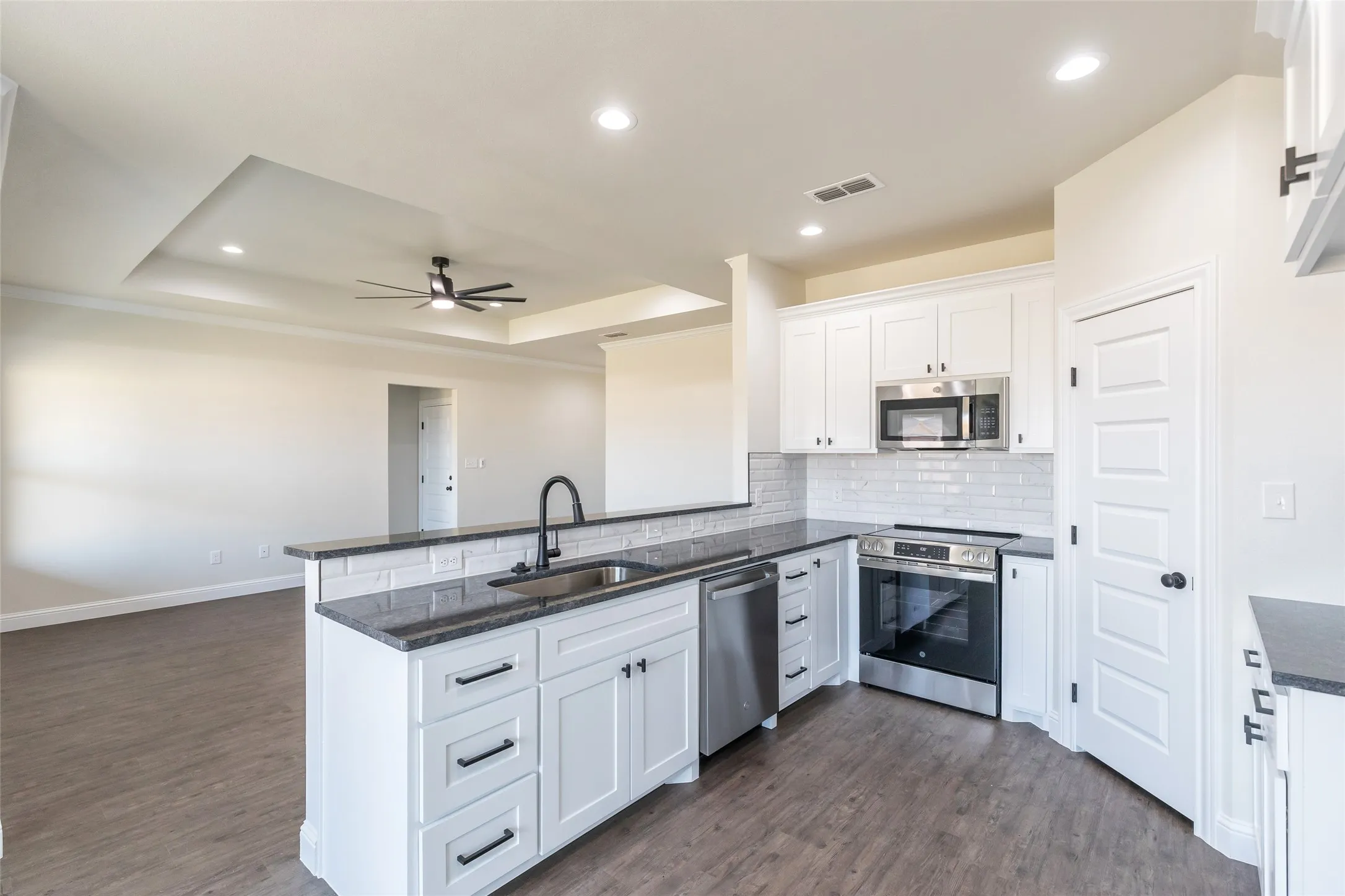 Modern kitchen featuring white shaker cabinetry, stainless steel appliances, dark countertops, a subway tile backsplash, and a large island with an undermount sink and matte black faucet