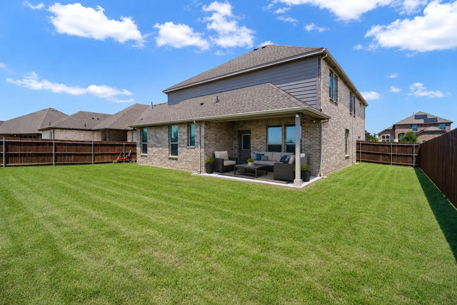 Expansive green lawn with a covered patio, brick exterior, dark brown privacy fencing, and multiple windows