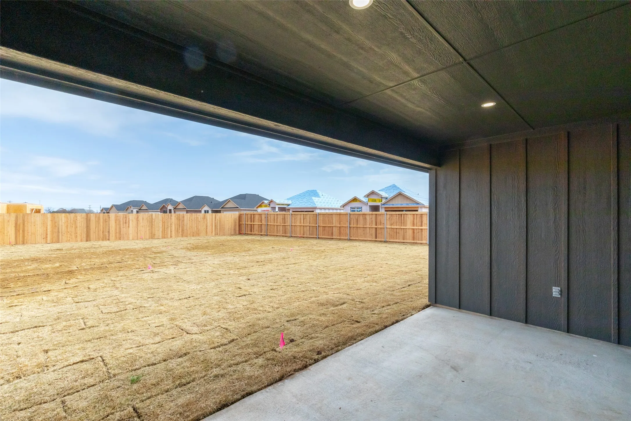 Covered concrete patio featuring recessed lighting, dark vertical panel siding, and a wooden privacy fence enclosing the backyard
