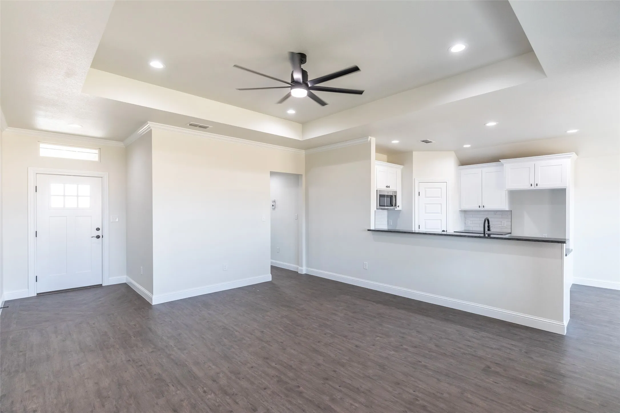 Open-concept interior featuring wood-finish flooring, a tray ceiling with recessed lighting and ceiling fan, and a kitchen bar with white cabinetry, a built-in microwave, and a dark countertop