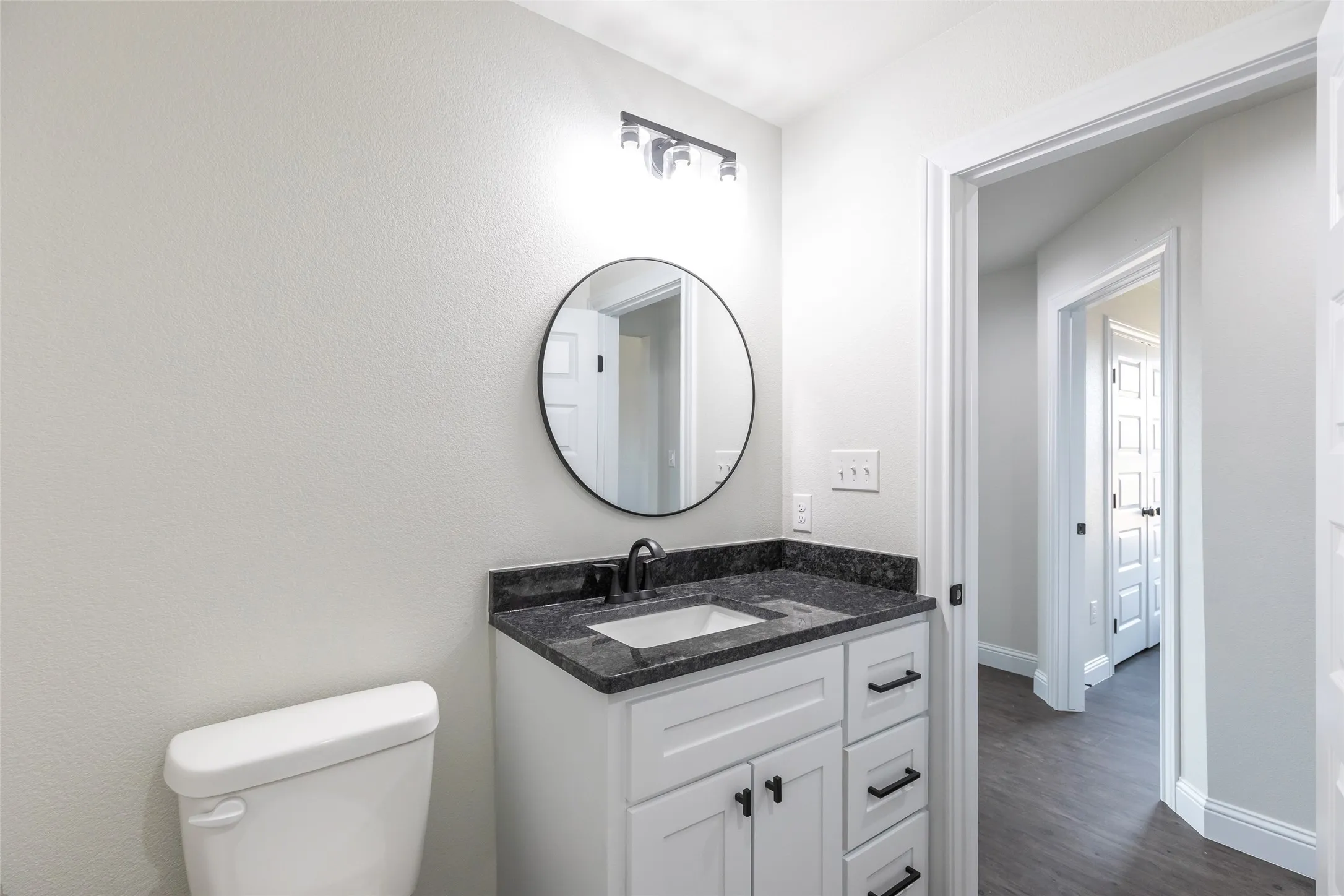 Bathroom featuring a white vanity with dark hardware, a dark speckled countertop, a rectangular undermount sink, a circular mirror, and dark wall-mounted lighting