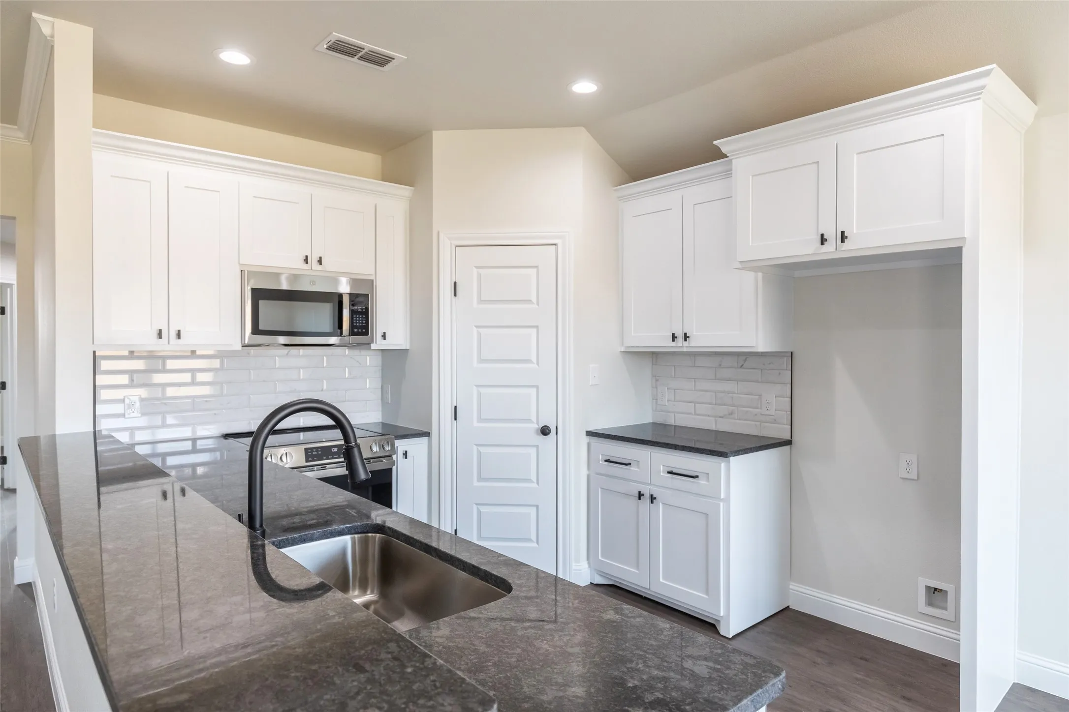 Modern kitchen featuring a large island with an undermount sink and matte black faucet, stainless steel microwave, white shaker-style cabinetry, and subway tile backsplash