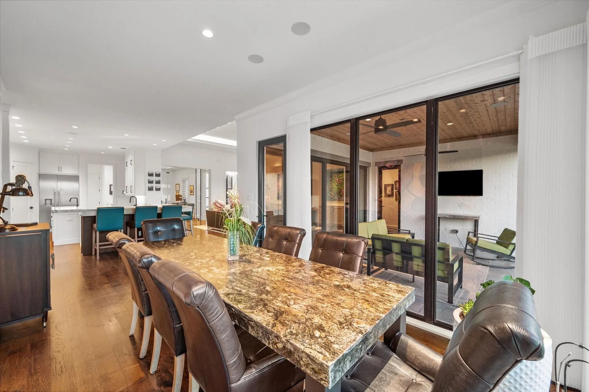 Open-concept interior featuring wood-finish flooring, a kitchen with stainless steel appliances and white cabinetry, a dining area with a stone-top table, and a covered outdoor living space with a wood-plank ceiling and a ceiling fan
