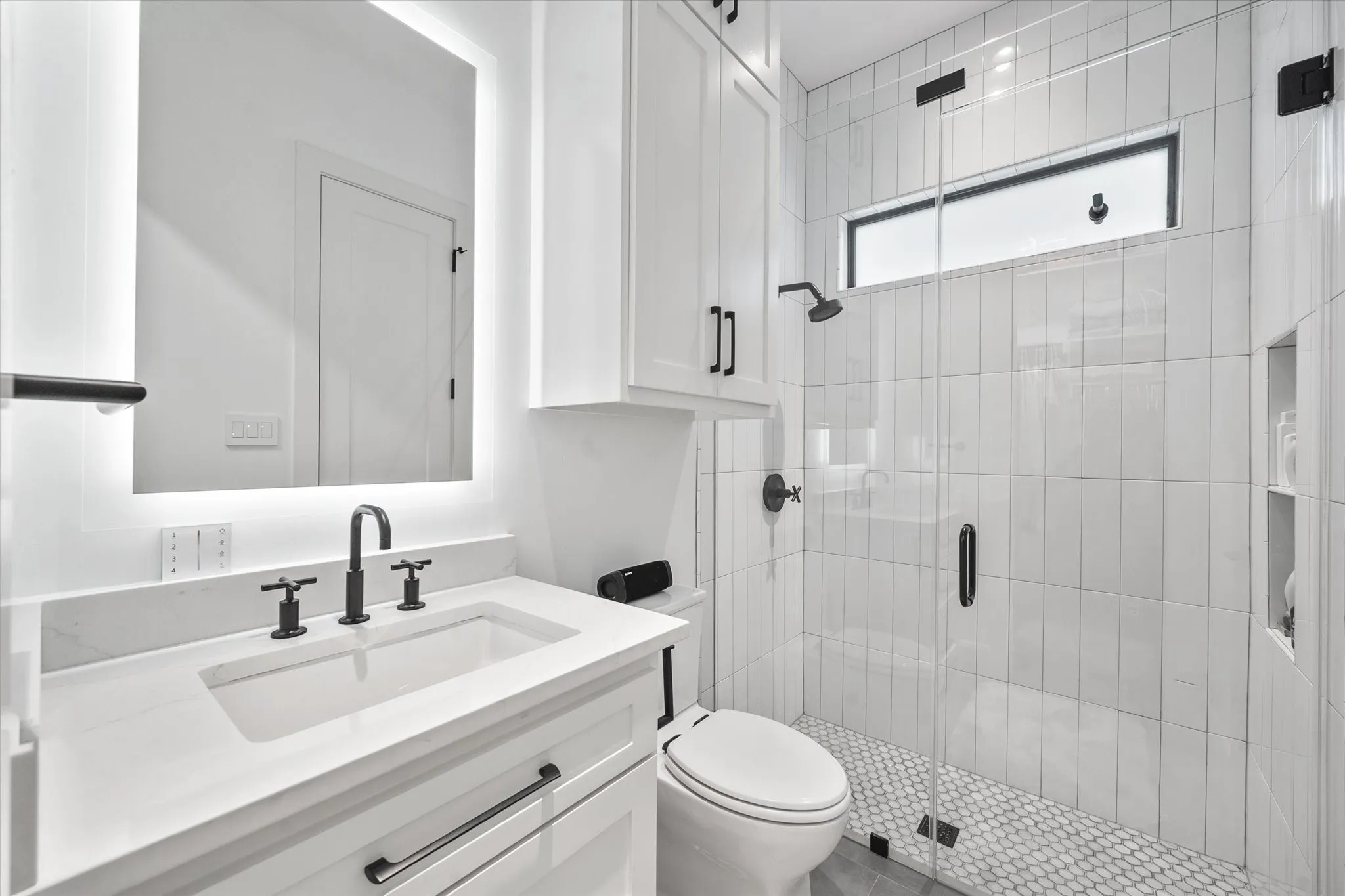 Modern bathroom featuring a frameless glass shower enclosure with white vertical tile, a penny tile shower floor, a floating vanity with a white countertop and integrated sink, a backlit mirror, and matte black fixtures