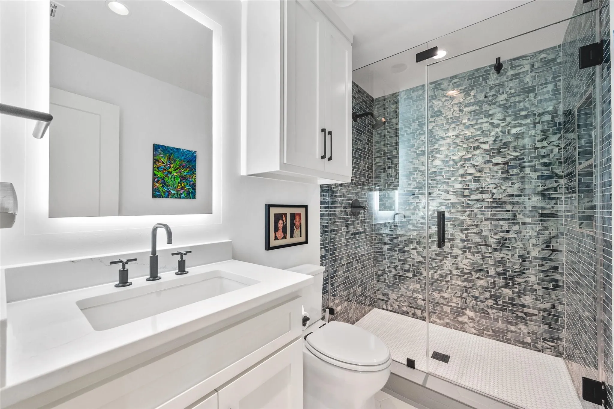 Modern bathroom featuring a frameless glass shower enclosure with mosaic tile surround, a vanity with a white countertop and rectangular undermount sink, a backlit mirror, and matte black plumbing fixtures