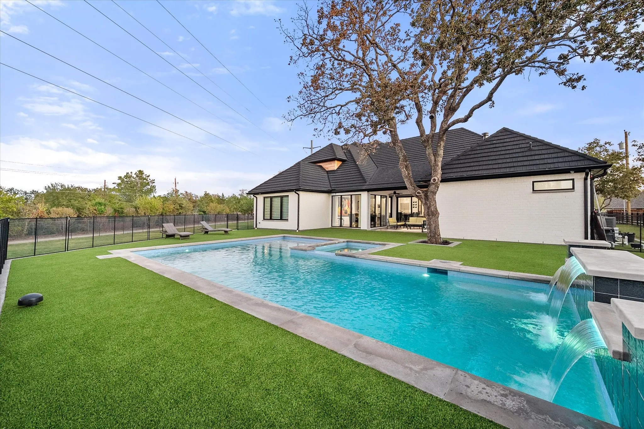 Expansive backyard featuring a rectangular swimming pool with integrated spa and waterfall features, surrounded by artificial turf and a black metal fence