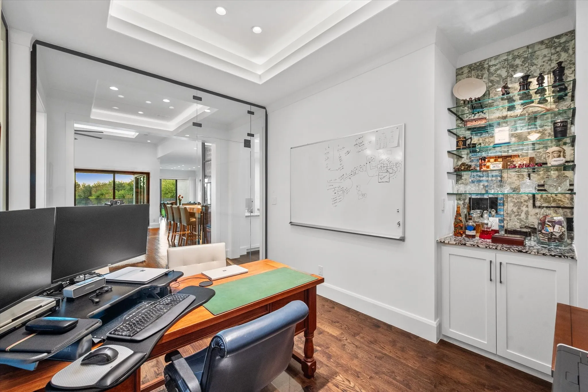 Dedicated workspace featuring glass panel walls, wood-finish flooring, a tray ceiling with recessed lighting, a built-in wet bar with a stone countertop, and custom shelving