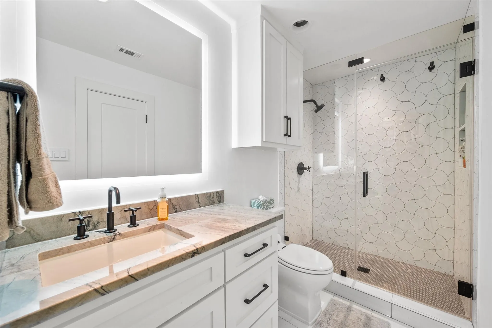 Bathroom featuring a vanity with a stone countertop and integrated sink, matte black hardware, a back-lit rectangular mirror, and white cabinetry