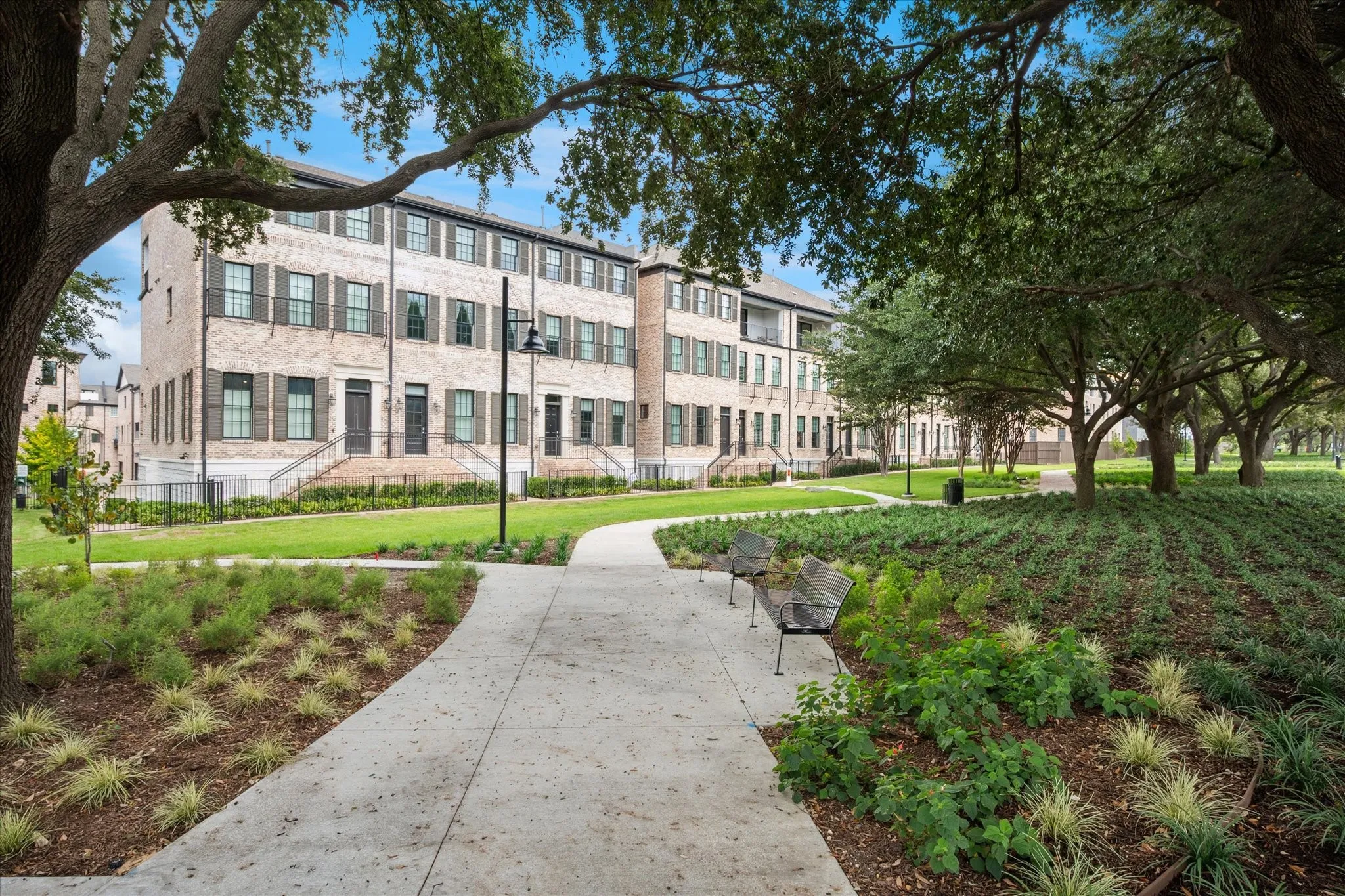 Brick facade townhomes featuring dark shutters and individual front entries, bordering a landscaped green space with mature trees