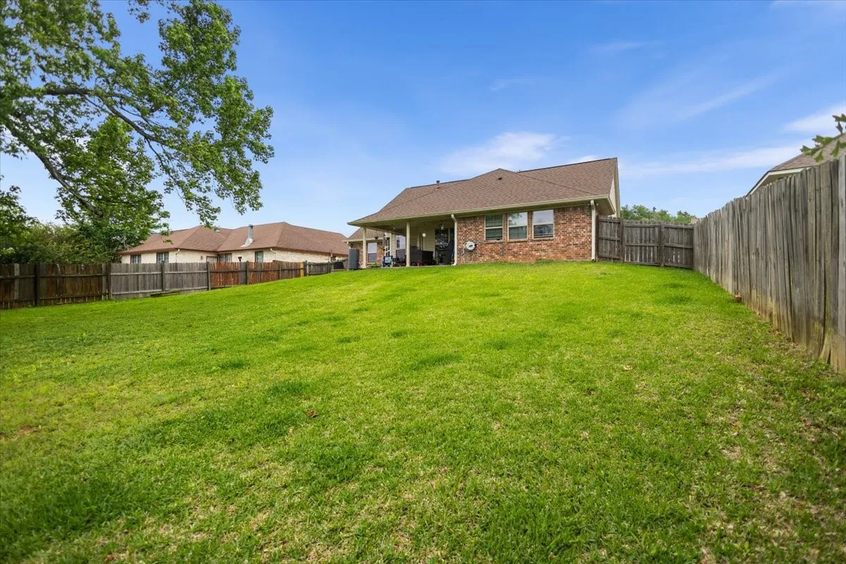 Expansive backyard featuring a lush green lawn, full privacy fencing, a covered patio, and a brick exterior