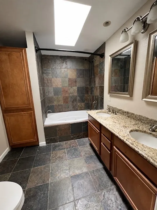 Skylight feature, tile flooring, dual vanity with undermount sinks, stone-look countertop, and a built-in soaking tub with tile surround