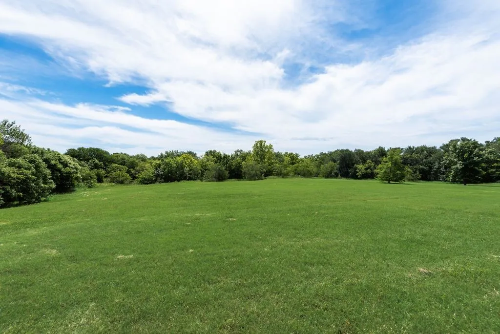 Expansive green space featuring a large grassy area, mature trees bordering the perimeter, and a clear sky with scattered clouds